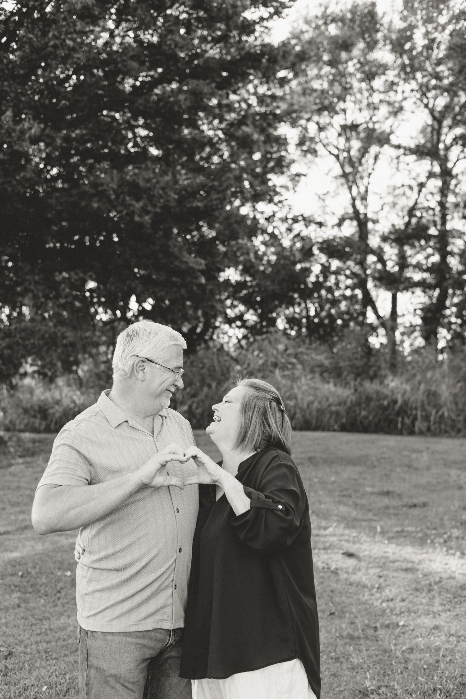 Black and white photo of a couple making a heart shape with their hands while smiling at each other outdoors.