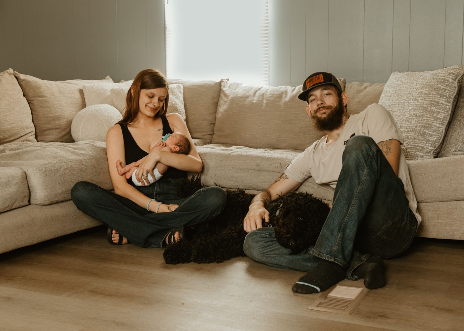 New parents sit on floor against beige sofa, mother nursing newborn baby while father relaxes beside them.