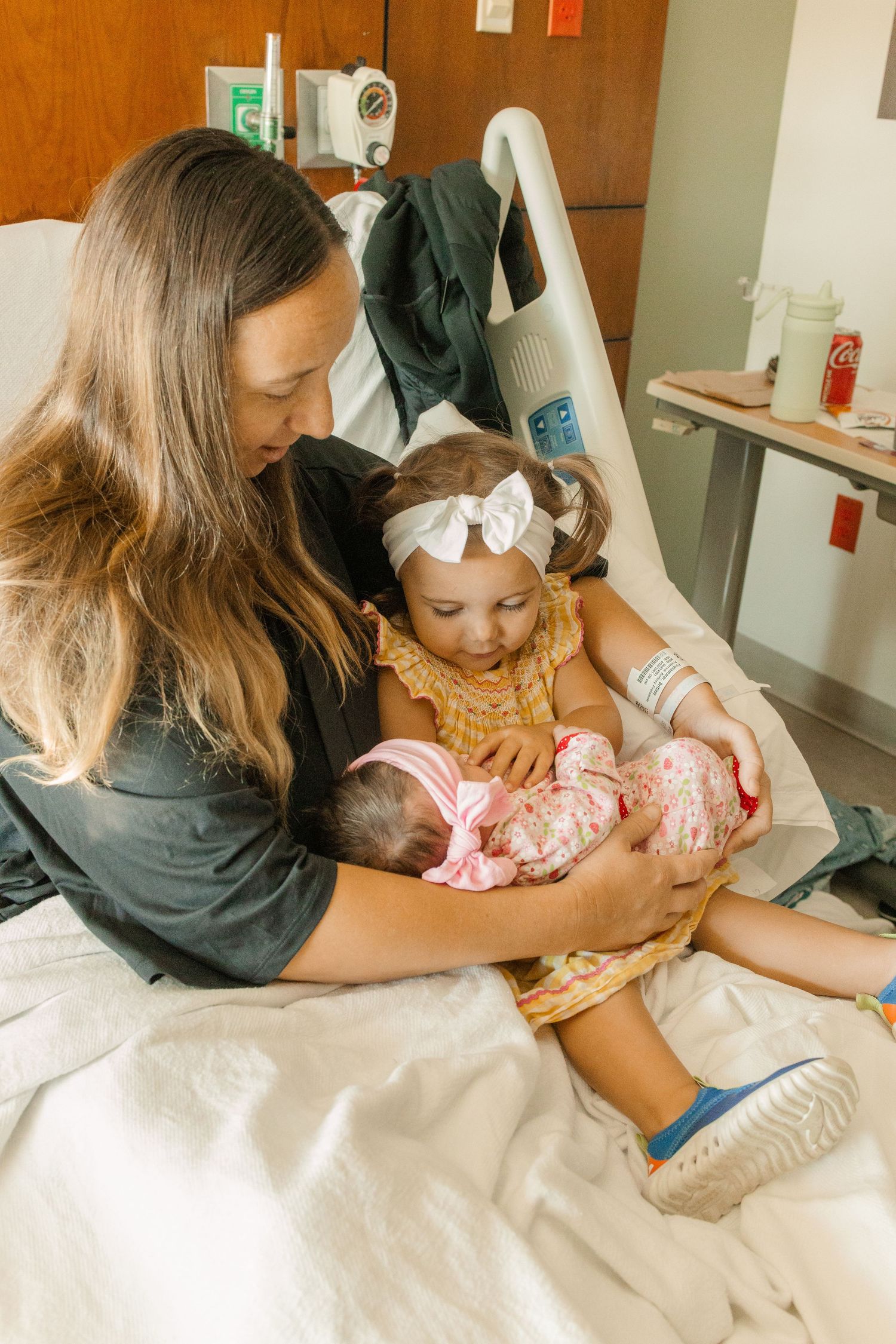 Mother sits in hospital bed holding newborn while toddler daughter looks on lovingly.