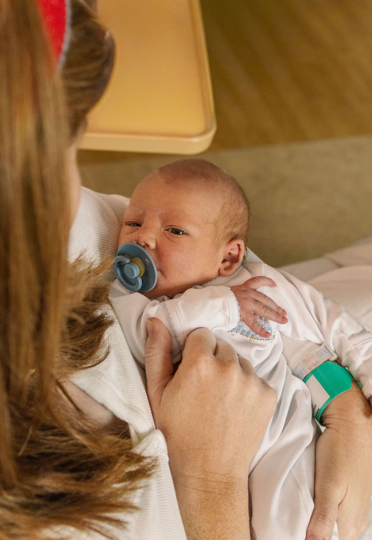 A newborn baby with a blue pacifier being held by a mother wearing a green hospital wristband.