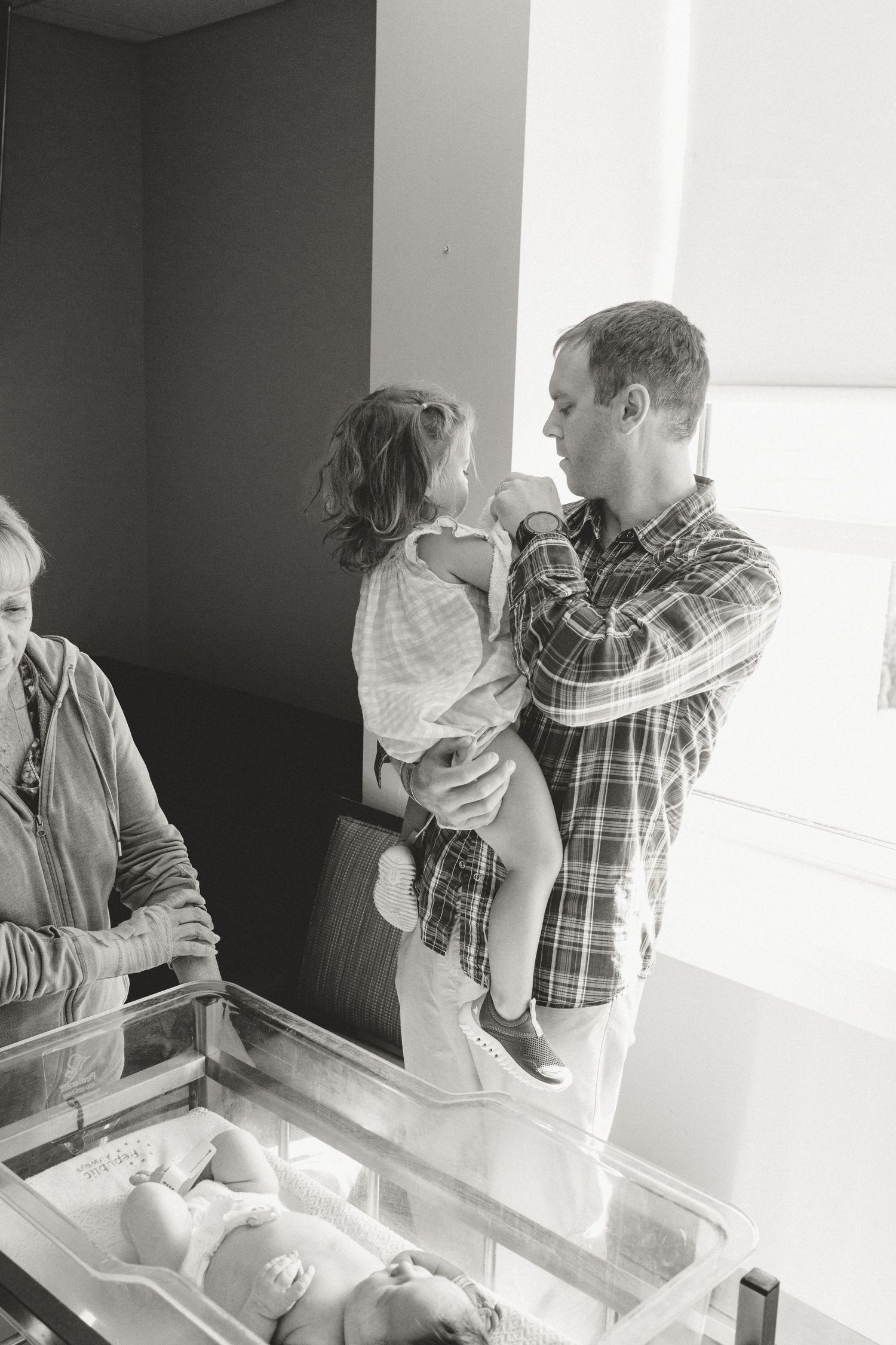 Father holds toddler daughter while viewing newborn sibling in hospital bassinet.