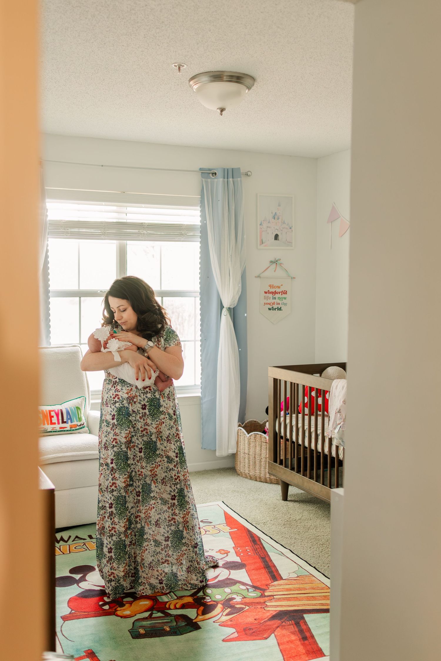 A brunette mother in a long floral dress holds her newborn in a decorated nursery by a bright window.