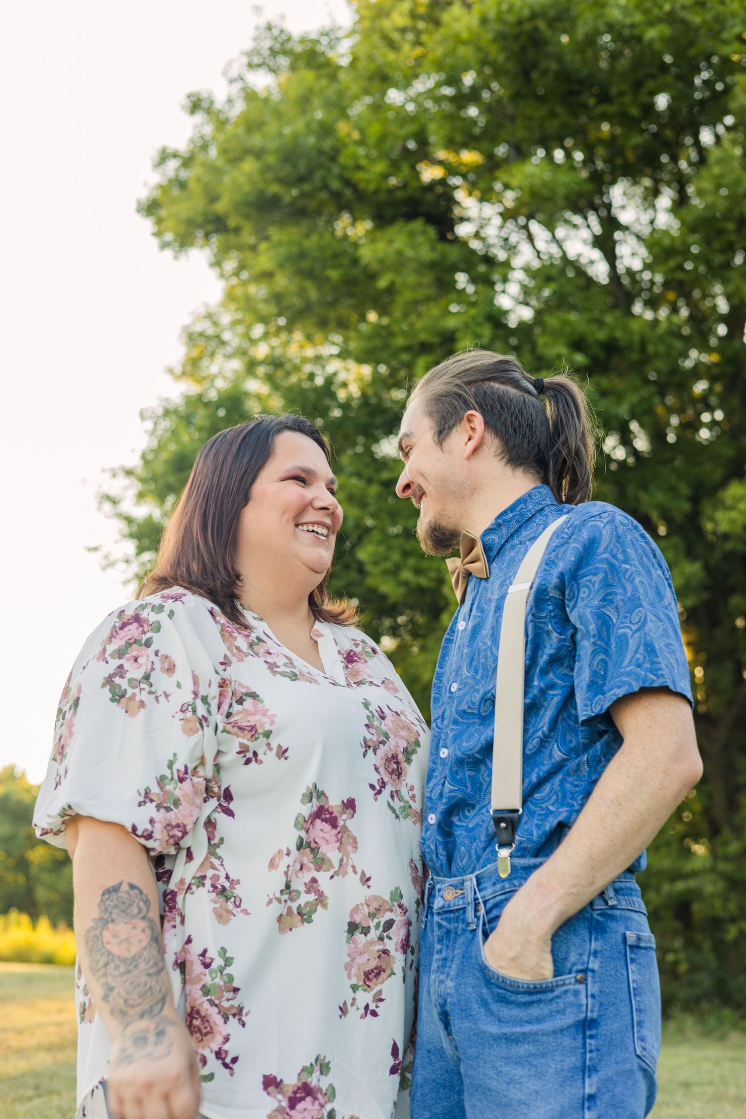 Happy couple laughing together outdoors at golden hour, woman in floral dress, man in bow tie and suspenders.