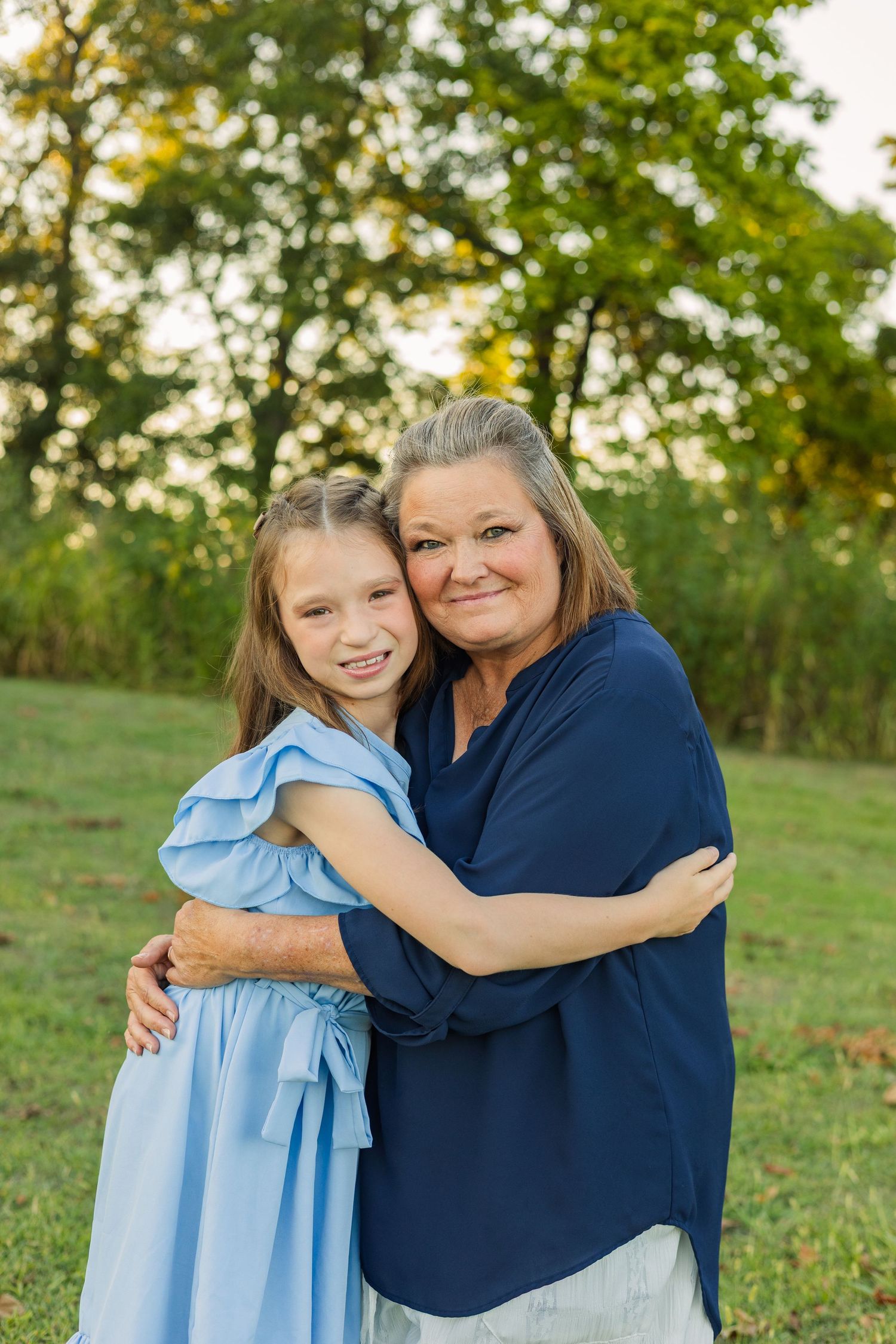 A smiling grandmother in navy blue hugging a young girl in a light blue dress outdoors.