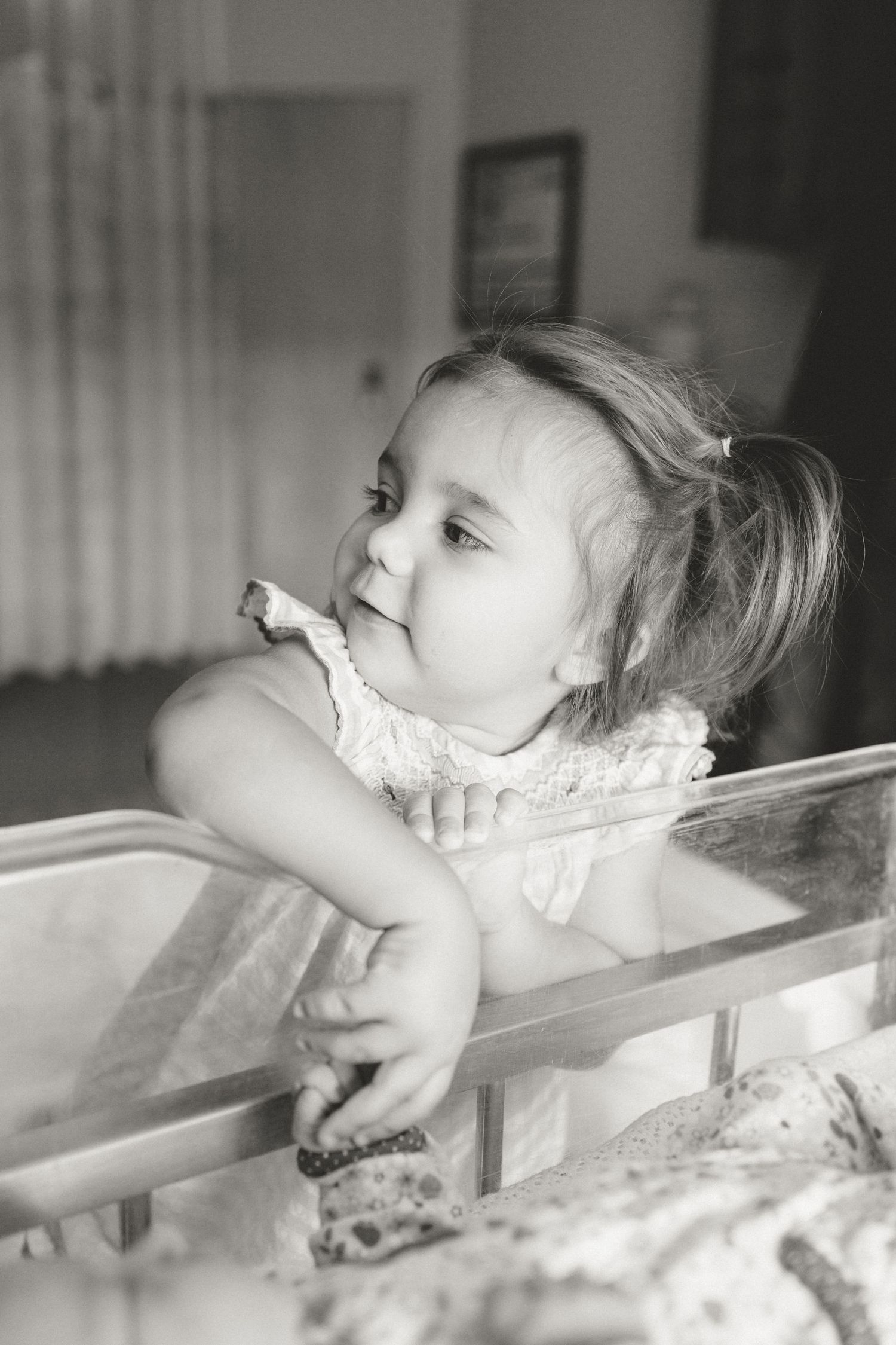 Black and white photo of toddler girl leaning on hospital newborn bassinet.