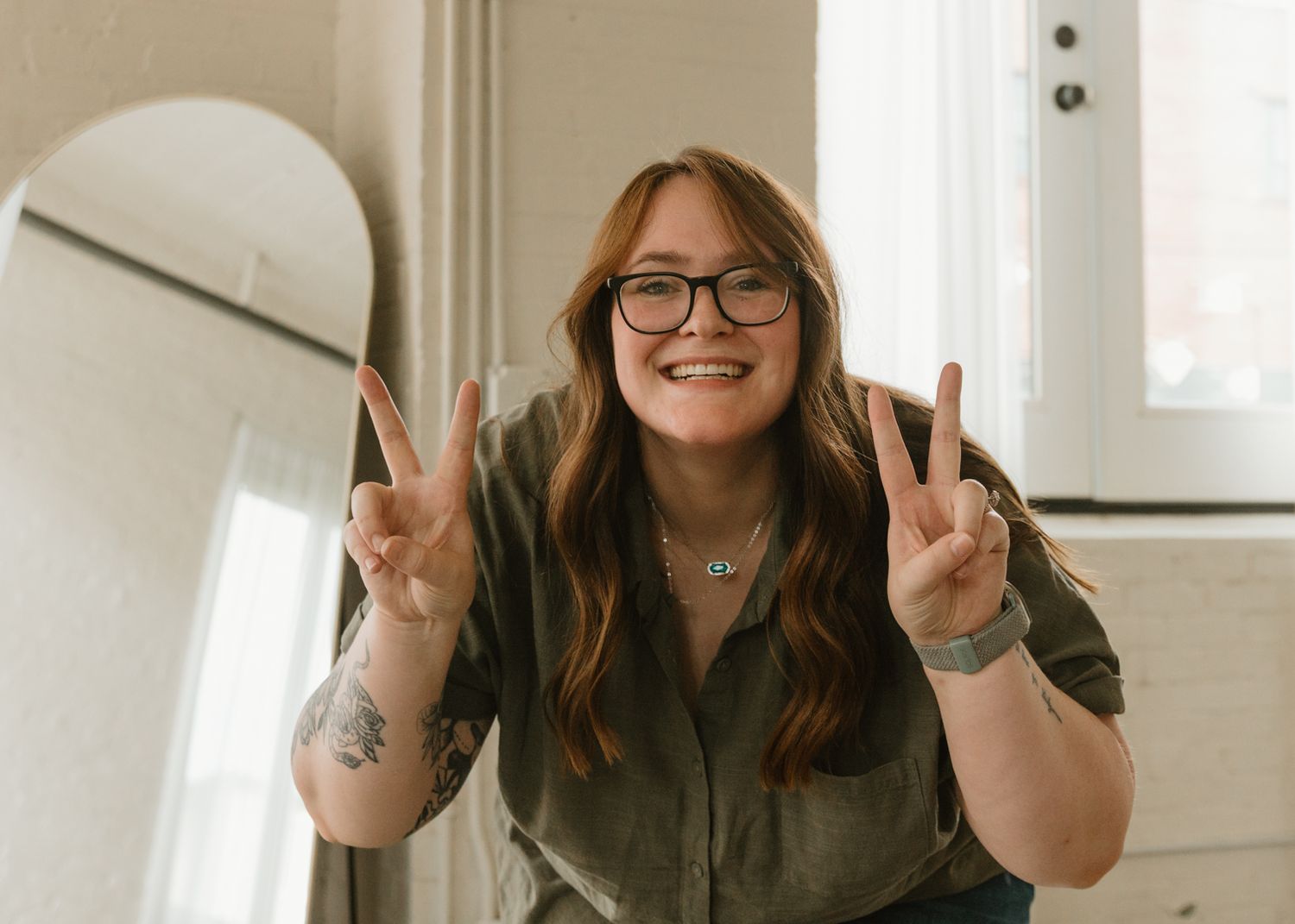 A smiling young woman with glasses and a tattoo gives two peace signs in a bright, airy room.