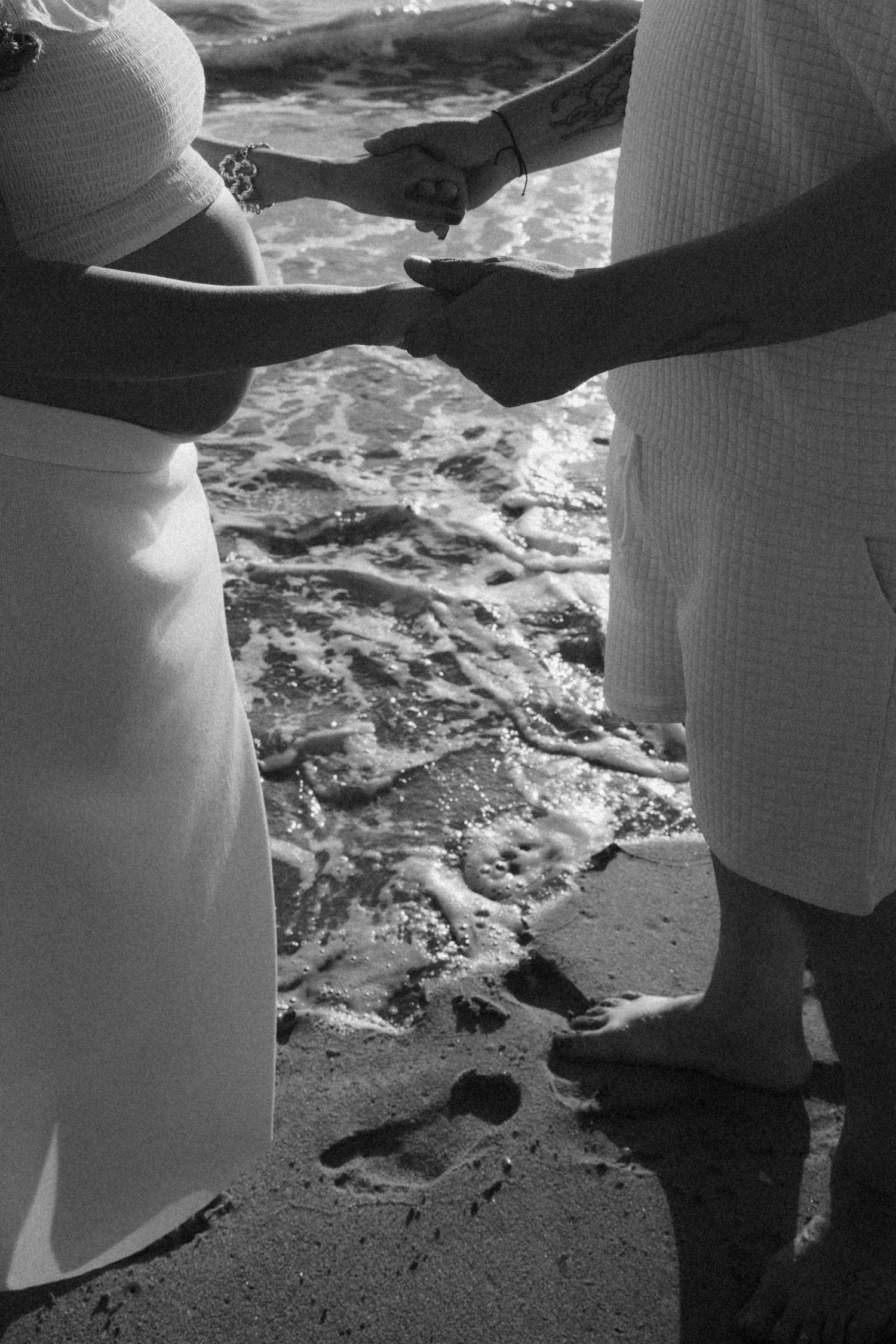 Black and white photo of pregnant couple holding hands at ocean shoreline with waves.