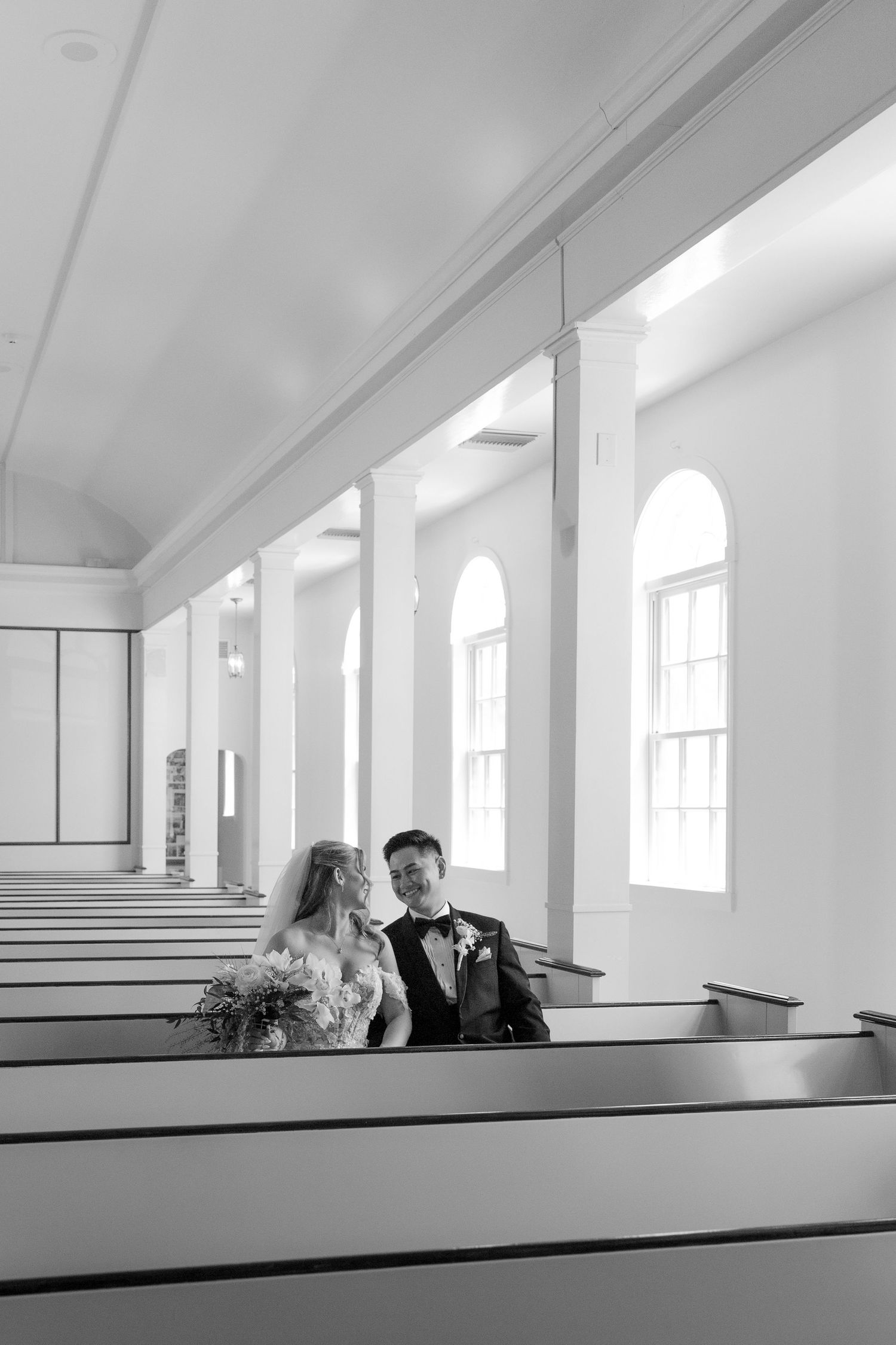 Black and white photo of bride and groom sitting together in elegant white church pews.