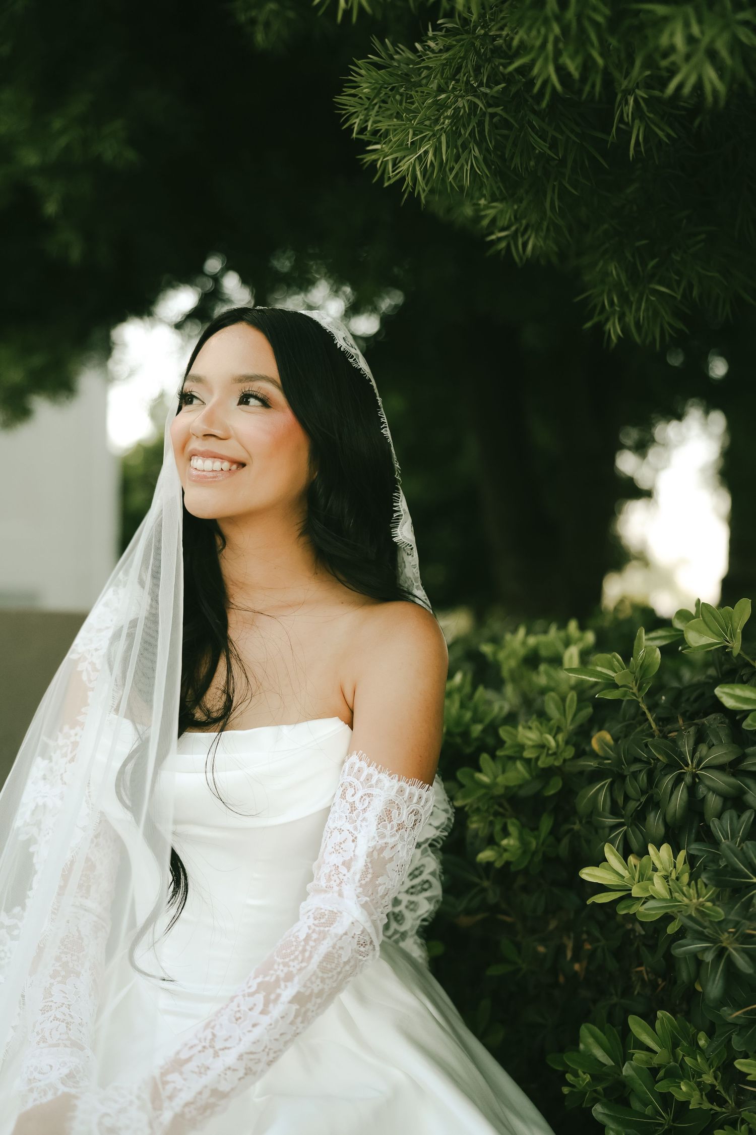 Smiling Asian bride in strapless white gown with lace veil poses beside lush green hedges outdoors.