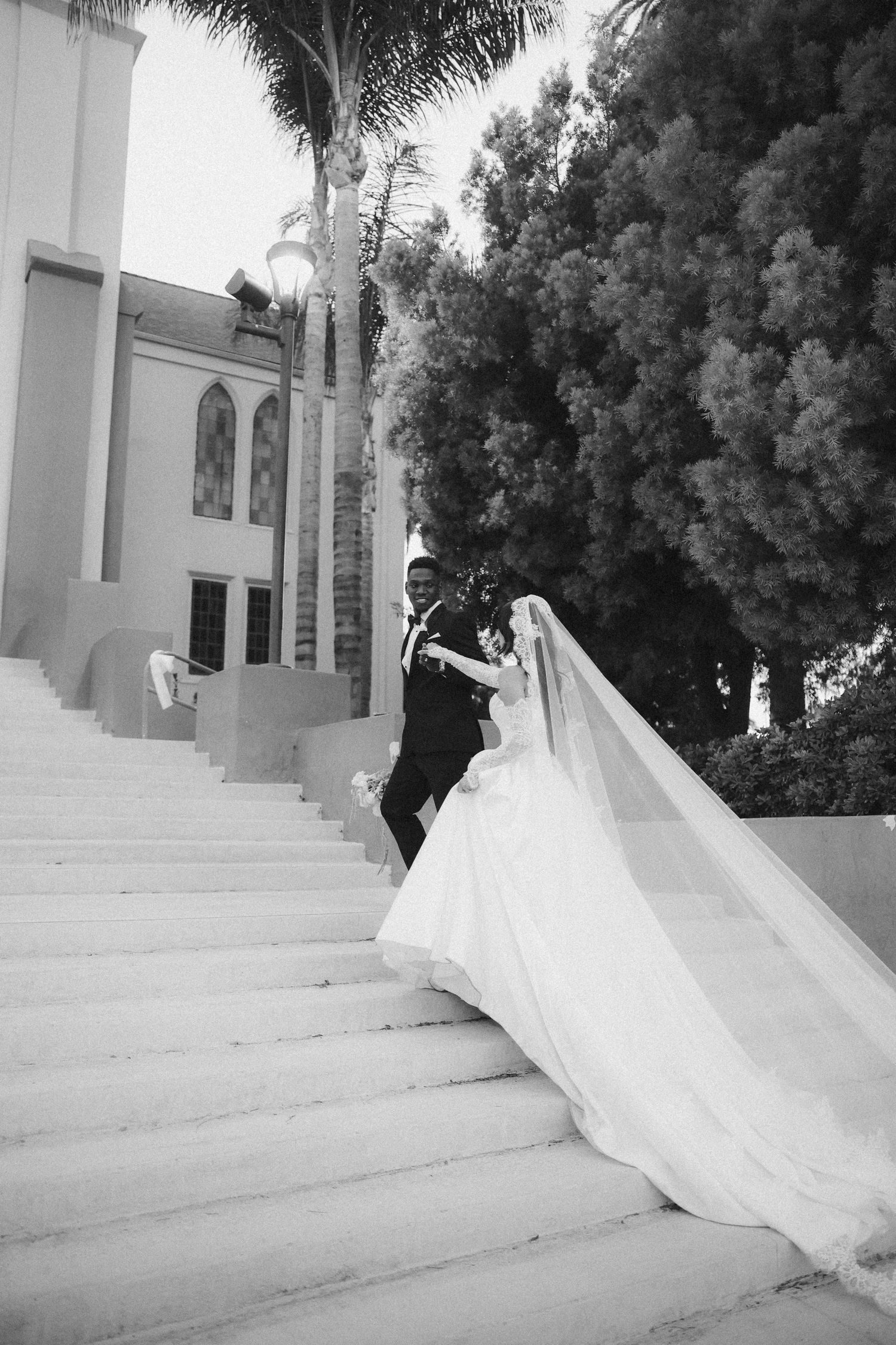 Black and white photo of bride and groom walking up grand white steps outside a church with palm trees.