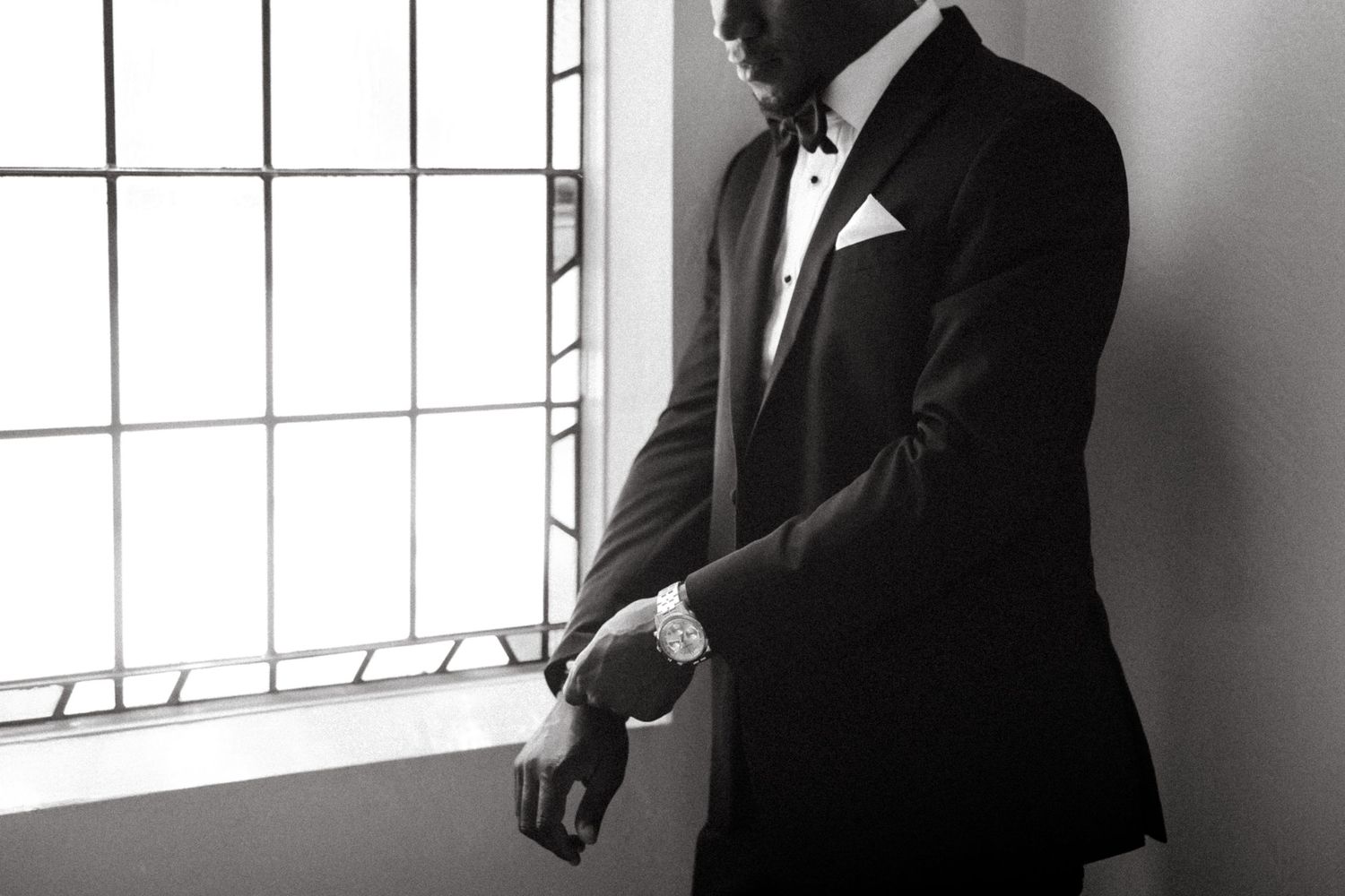 Elegant groom in black tuxedo adjusting watch by window in black and white wedding portrait.