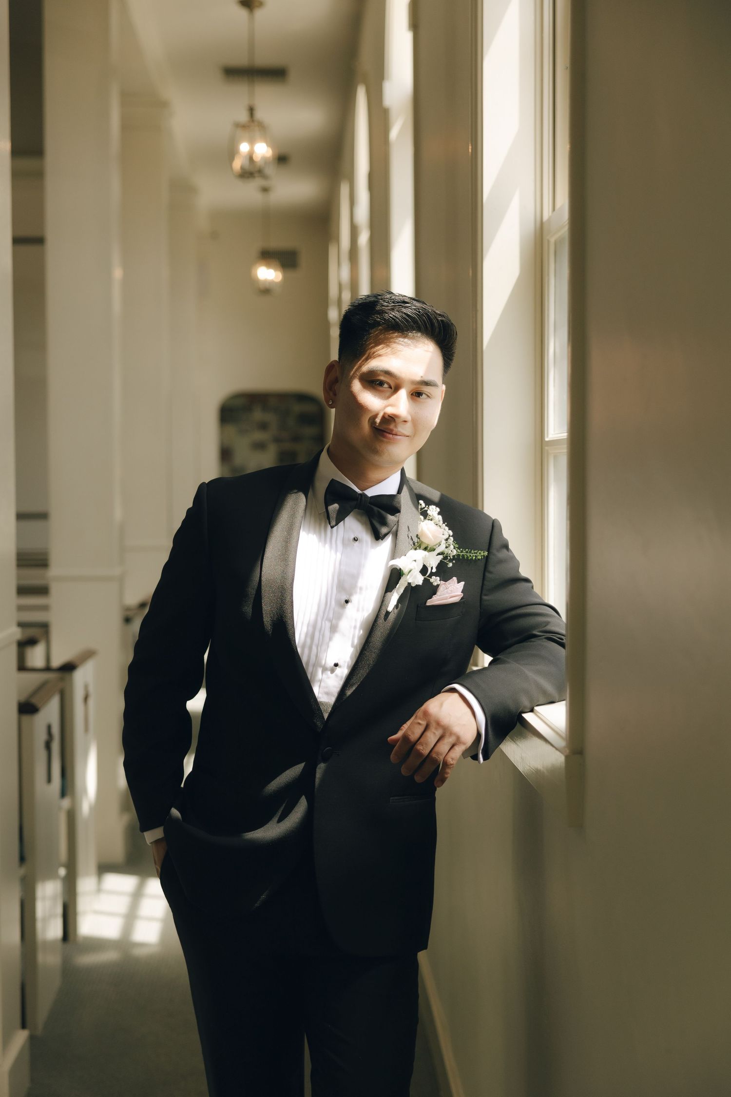 Dapper groom in black tuxedo with bow tie and boutonniere leans against window in elegant hallway.