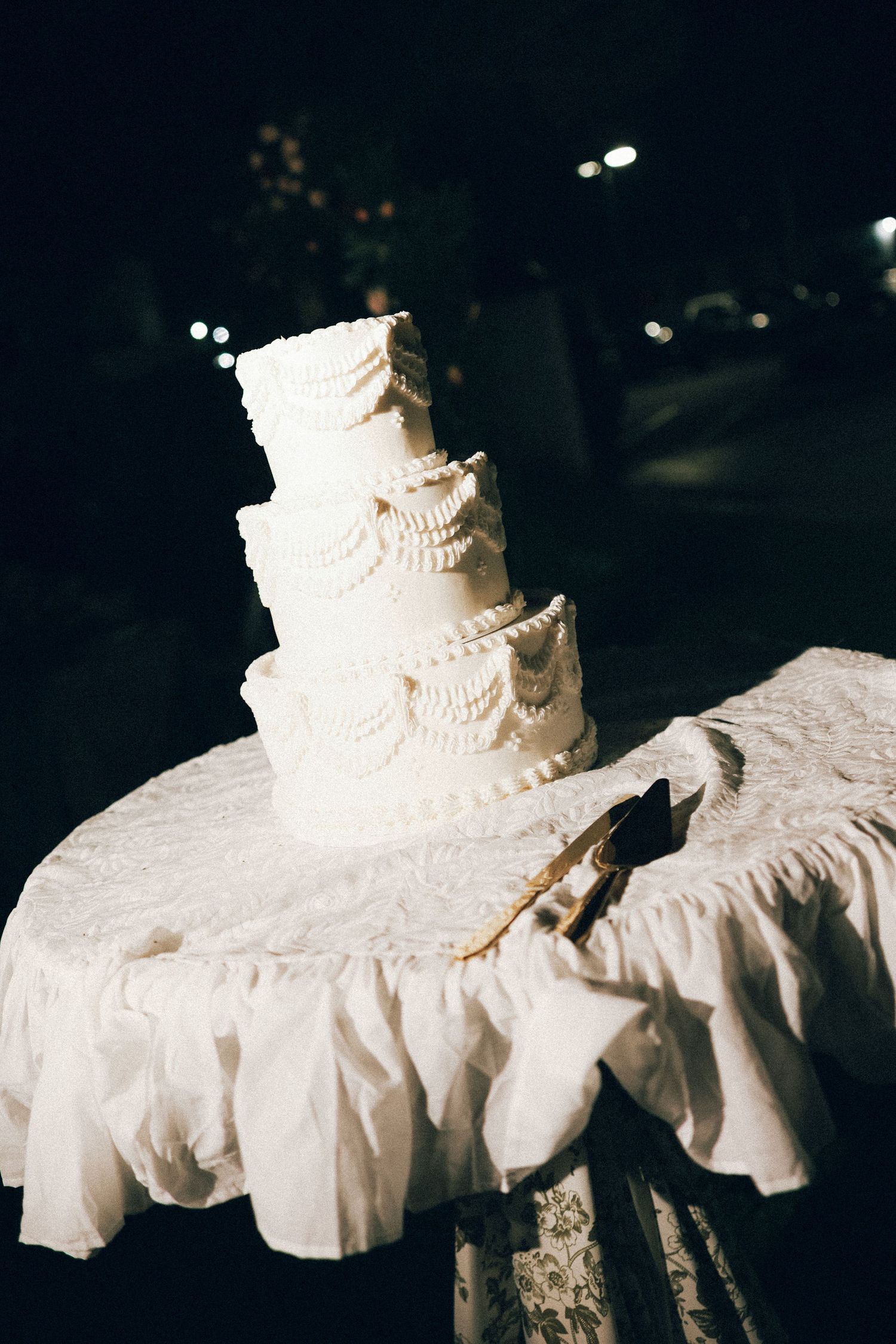 Elegant four-tier white wedding cake with ruffled frosting displayed on a draped table at a nighttime reception.