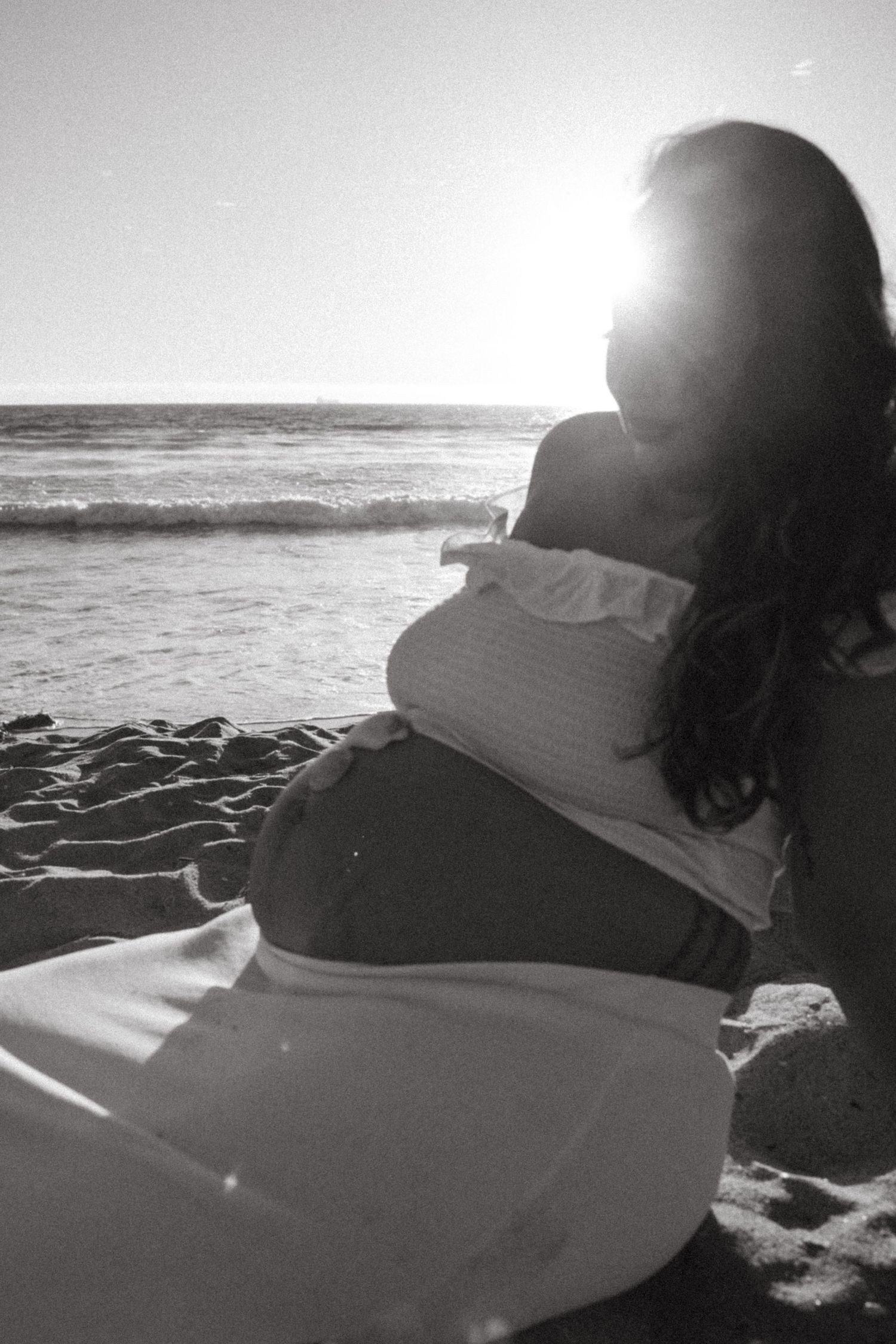 Black and white silhouette of pregnant woman sitting on beach with sun flare behind her.