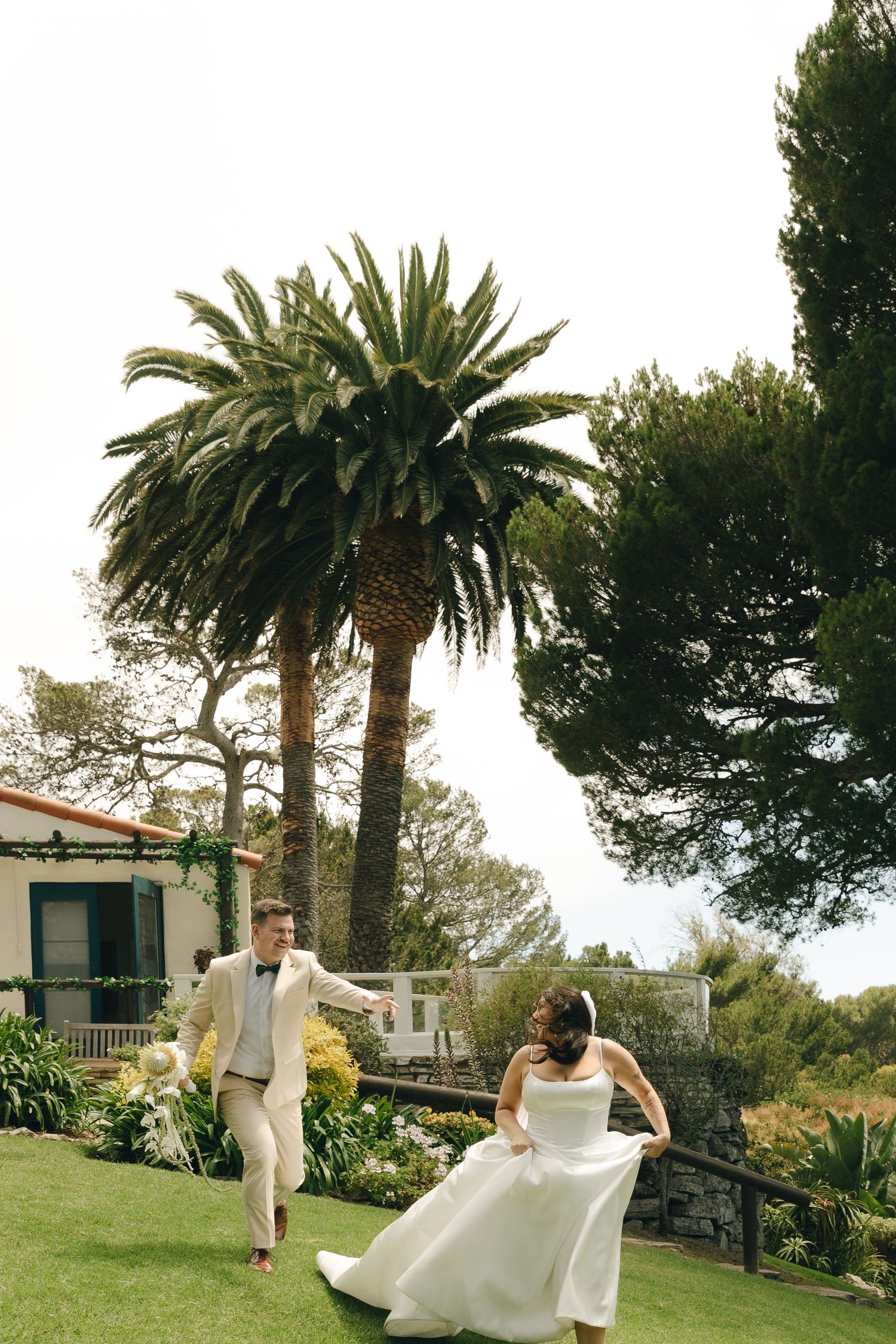 Bride and groom playfully run across lush garden lawn with tropical palm trees and Mediterranean villa in background.