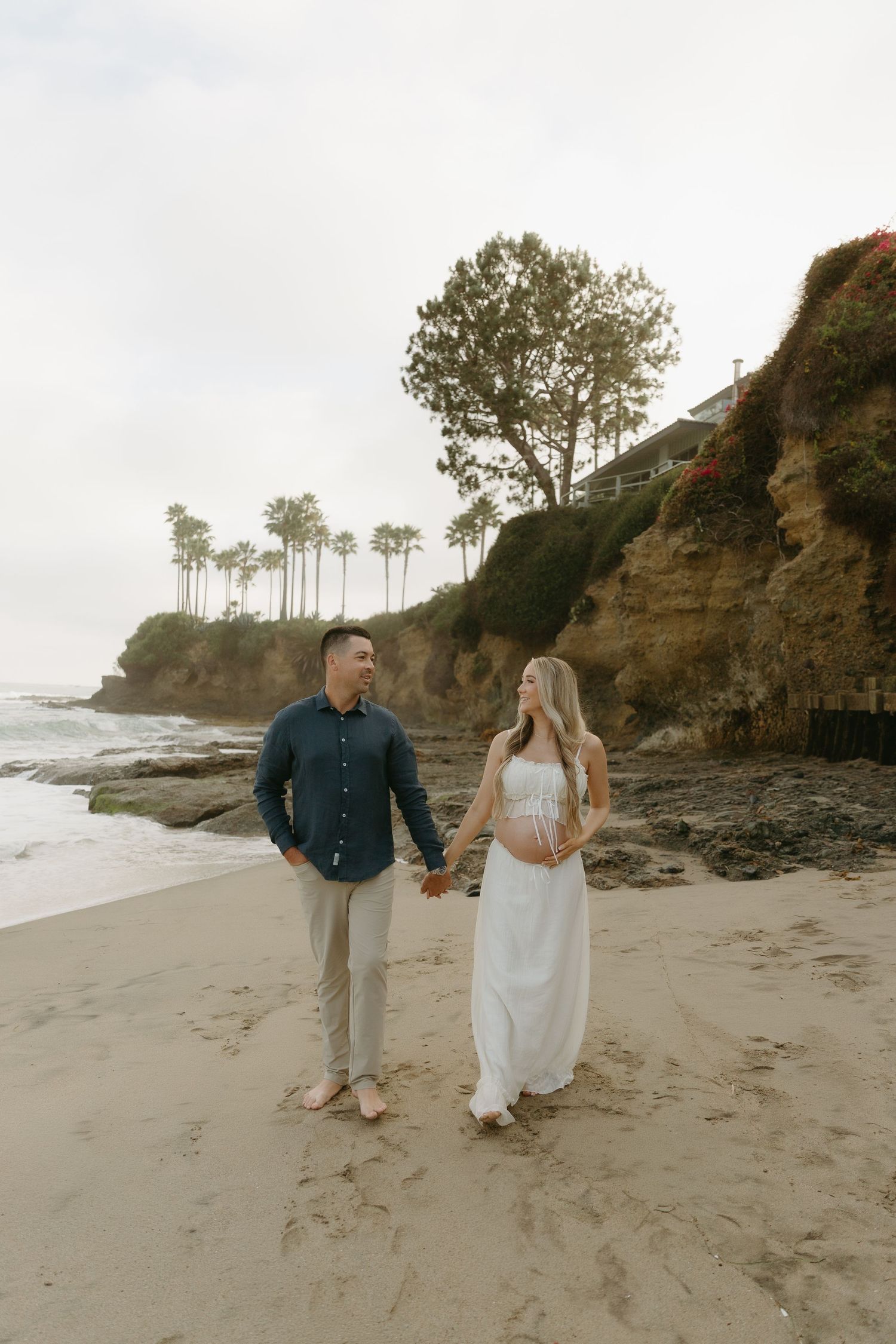 Expecting couple holding hands and walking on sandy beach with rocky cliffs and palm trees in background.