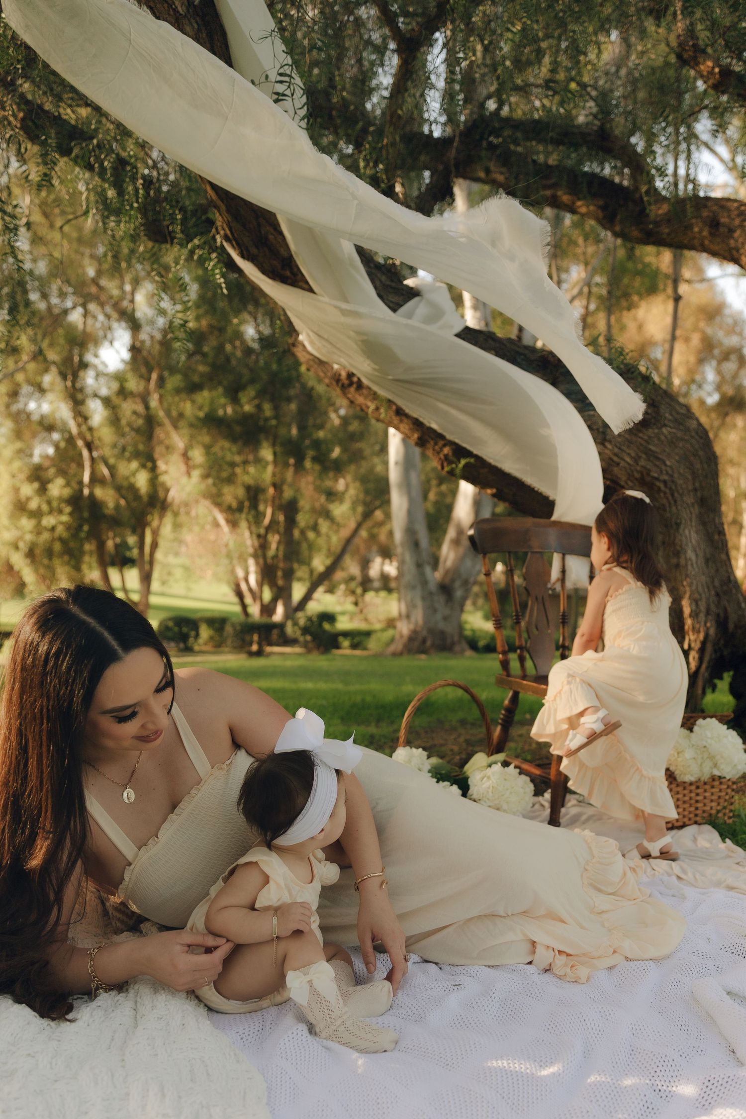 Mother in cream dress nursing baby on picnic blanket while toddler plays under large tree.