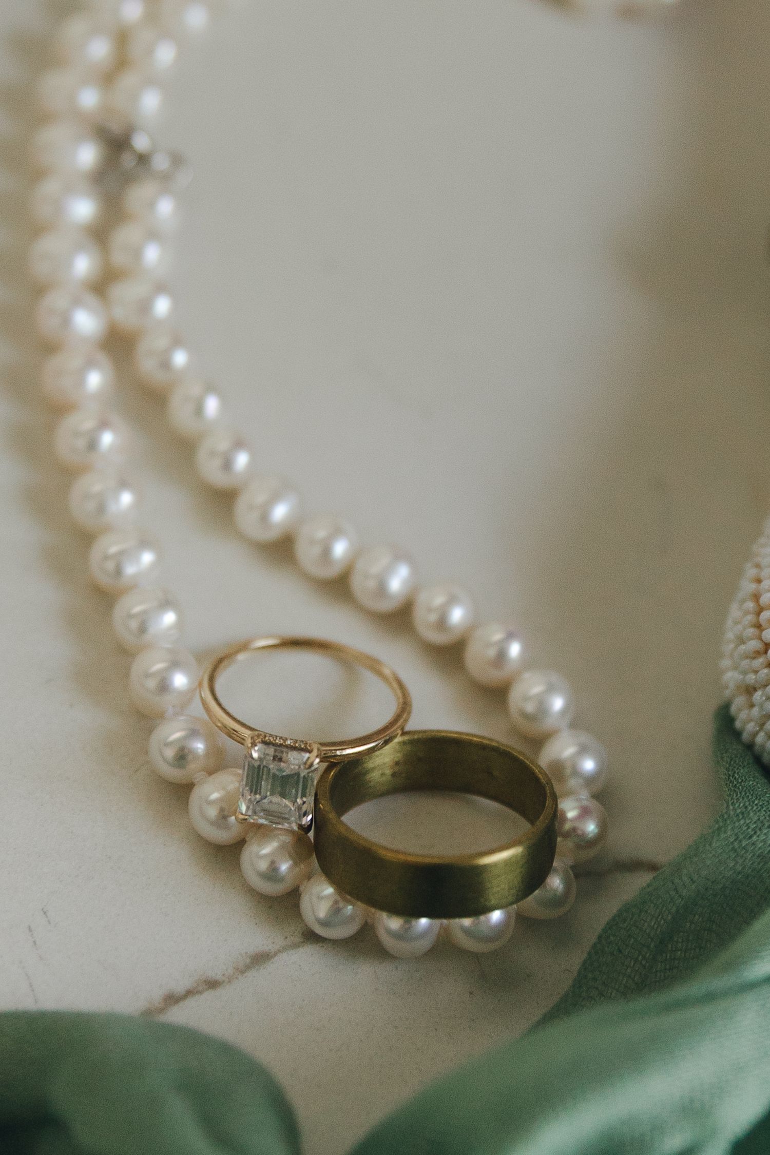Gold engagement ring and wedding band resting on pearl necklace on white marble surface.