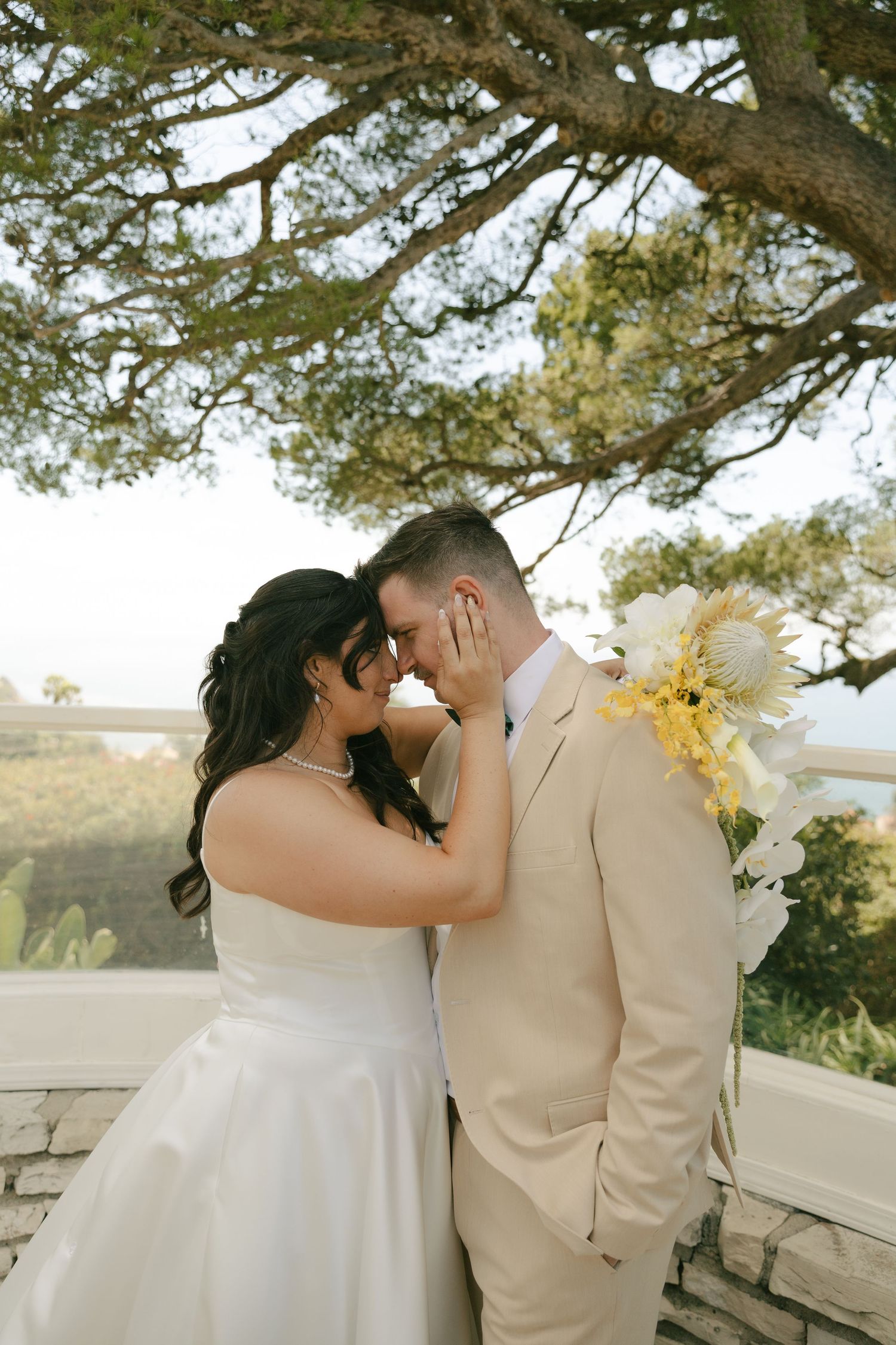 Bride and groom touch foreheads tenderly under a sprawling tree, groom wears beige suit with floral boutonniere.