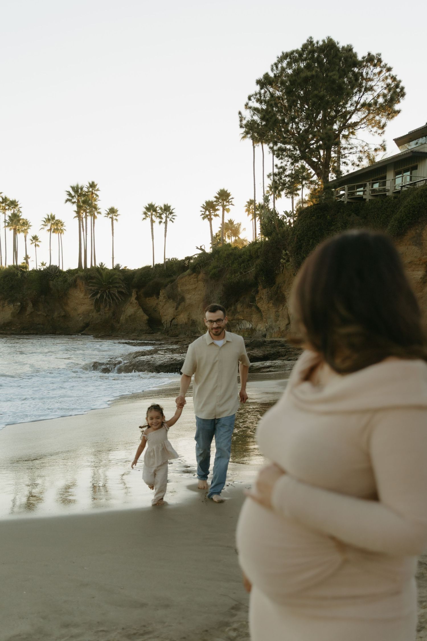 Father walks with toddler on sandy beach at sunset while pregnant mother stands in foreground.