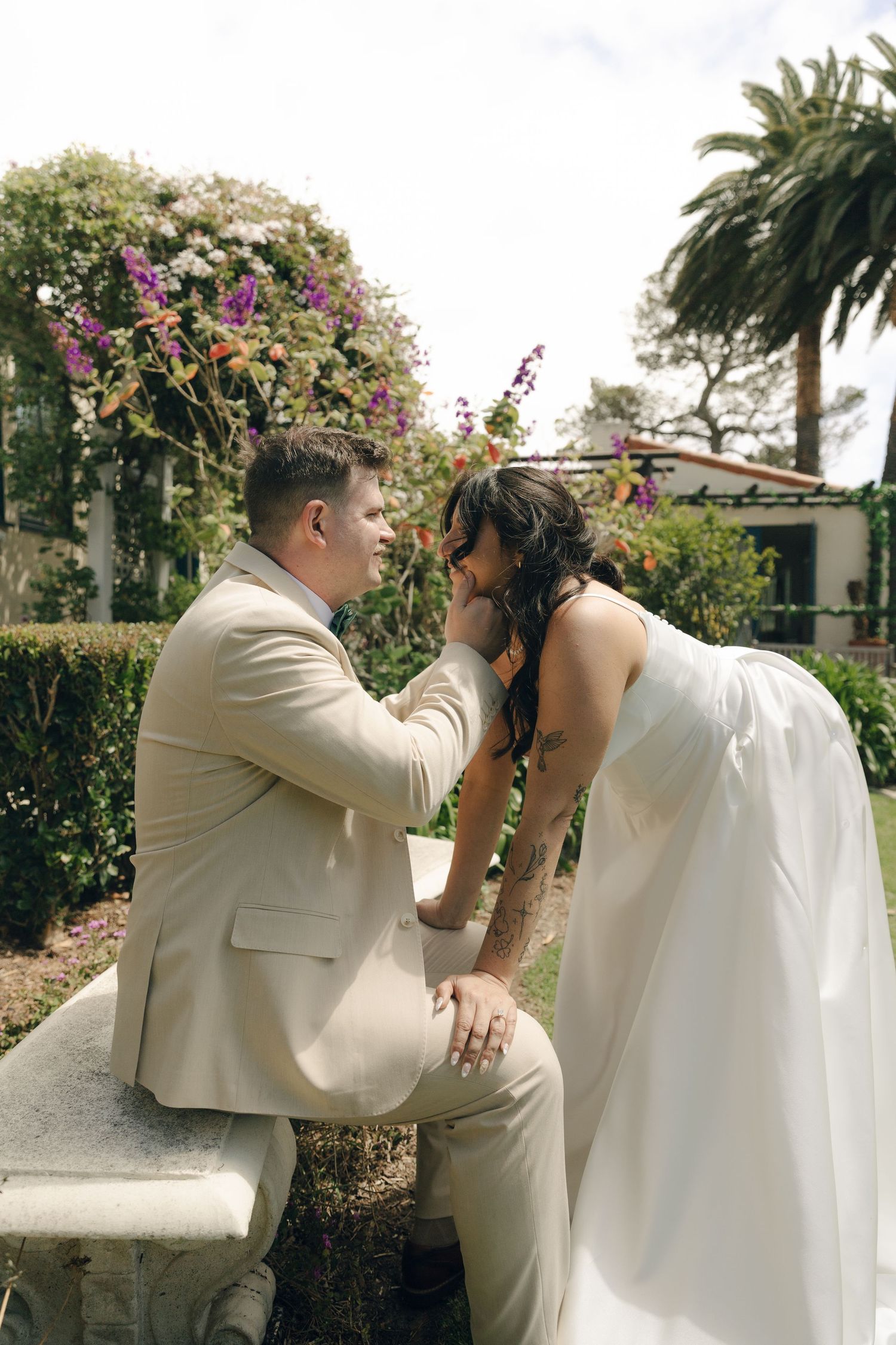 Groom in beige suit sits on stone bench holding brides hands in colorful garden with bougainvillea blooms behind them.