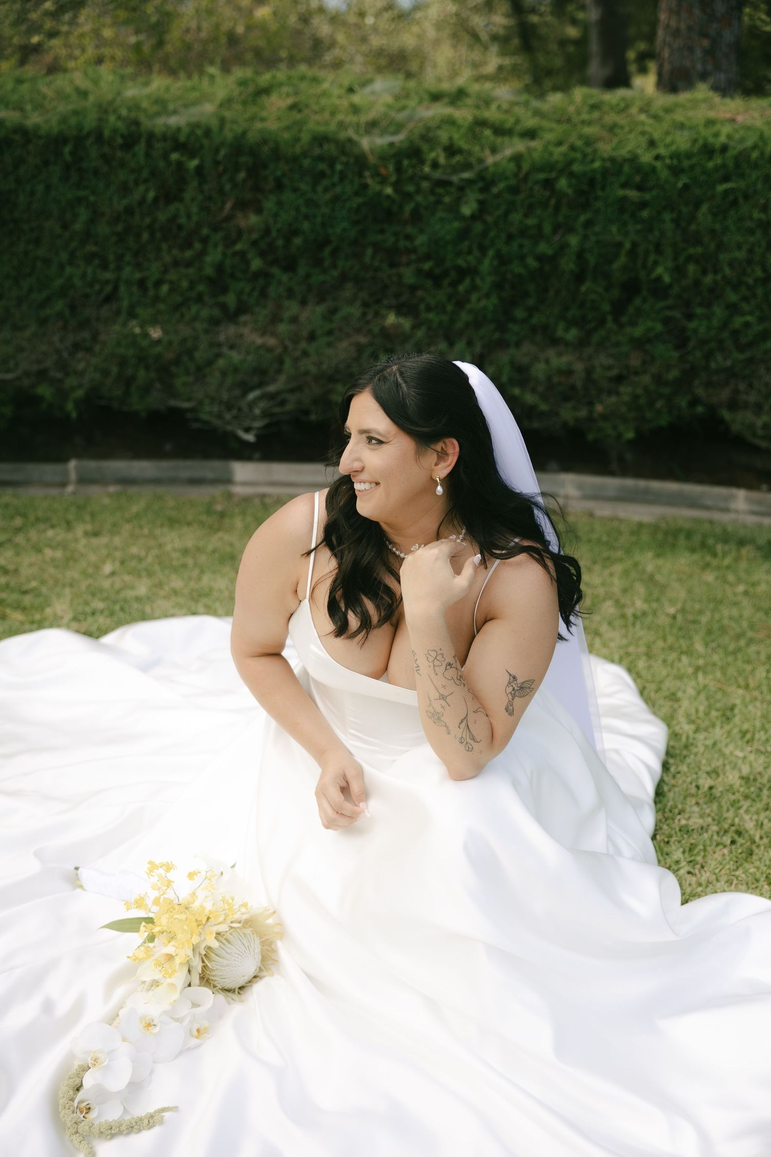 Smiling bride in white wedding dress sitting on lawn with floral bouquet in garden.