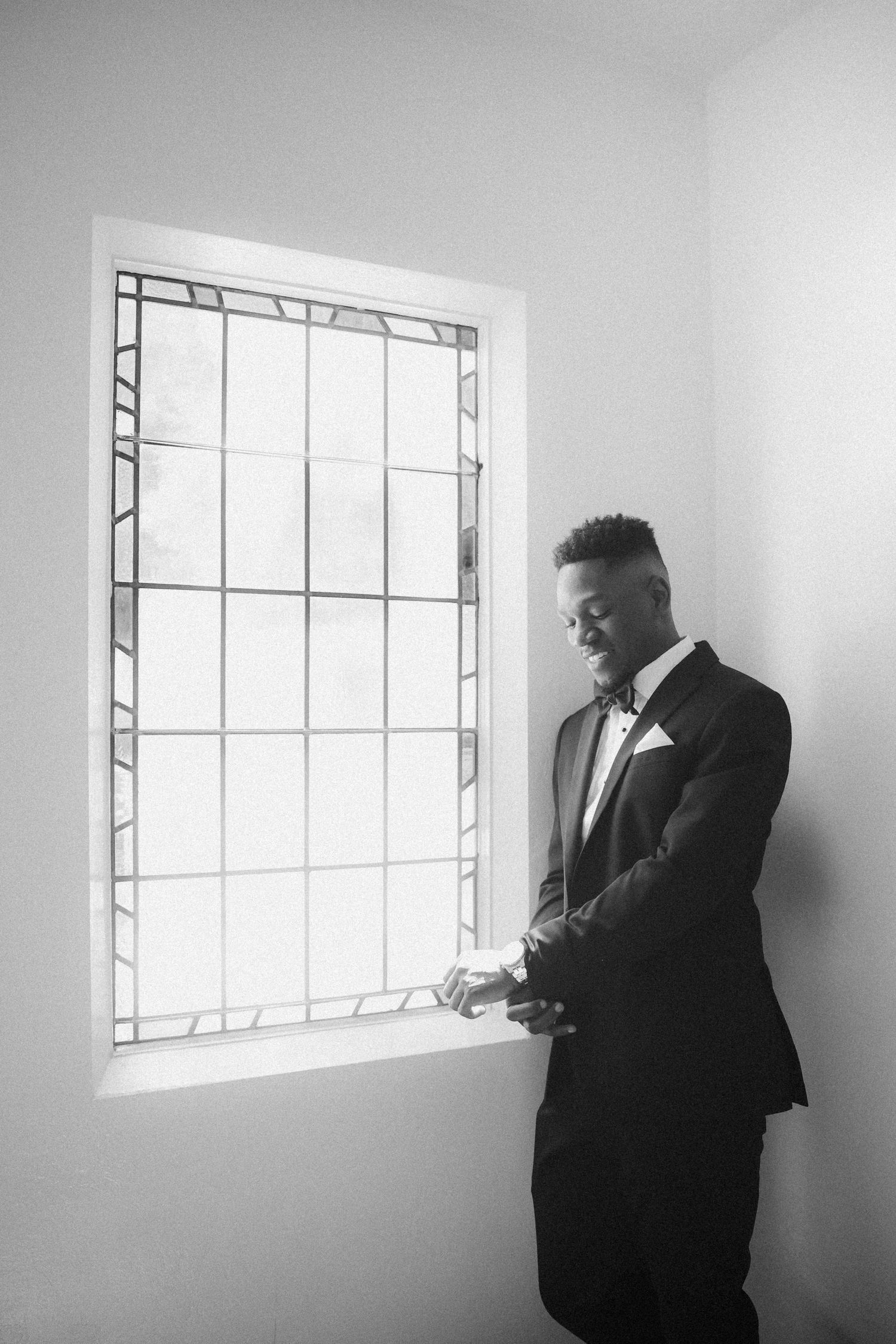 Handsome groom in black tuxedo standing by window adjusting cufflinks in black and white photo.