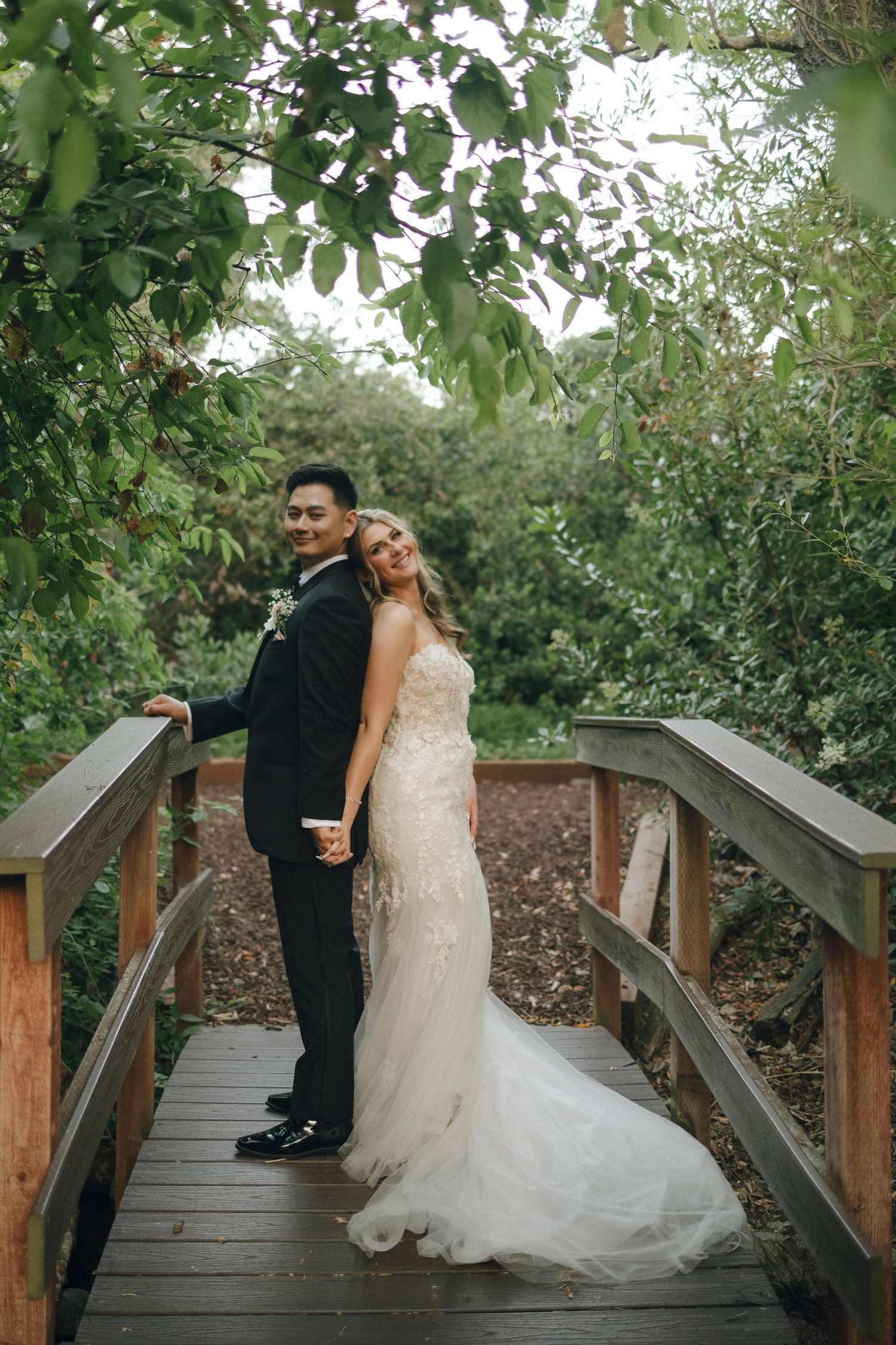 Bride and groom pose on rustic wooden bridge surrounded by lush green foliage on wedding day.
