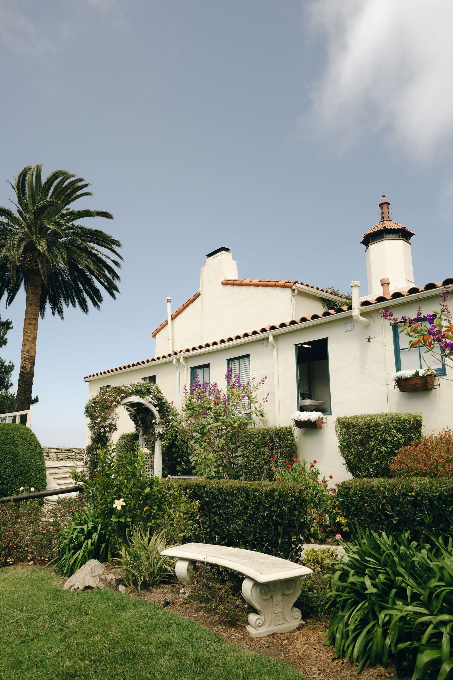 White Spanish-style villa with terracotta roof, palm trees, and stone bench in manicured garden.