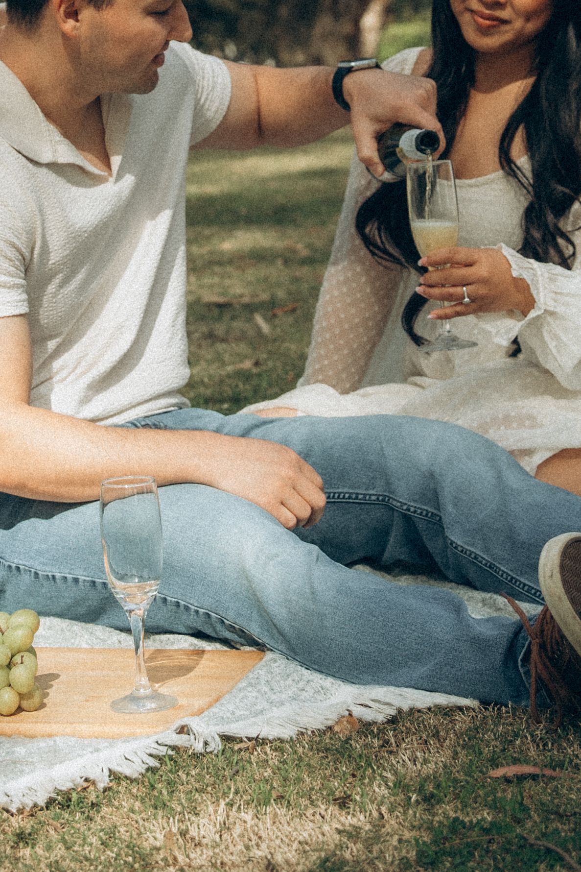 Couple enjoying a romantic picnic, pouring champagne into glasses with grapes on a wooden board outdoors.