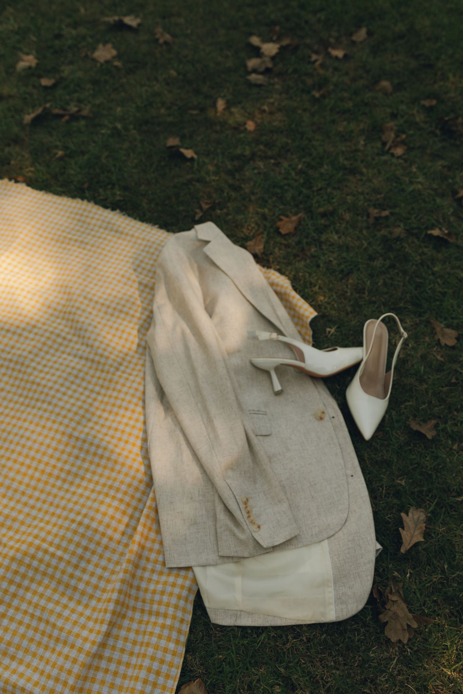 Gray blazer and white bridal heels resting on a yellow plaid blanket on grass with autumn leaves.