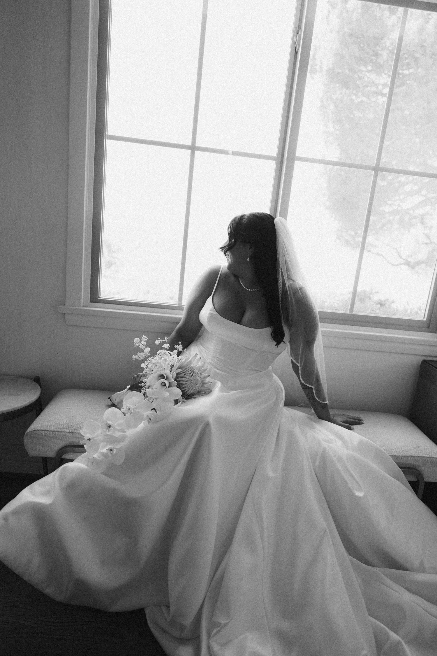 Black and white photo of a bride in a flowing white gown and veil, seated by a window holding a bouquet.