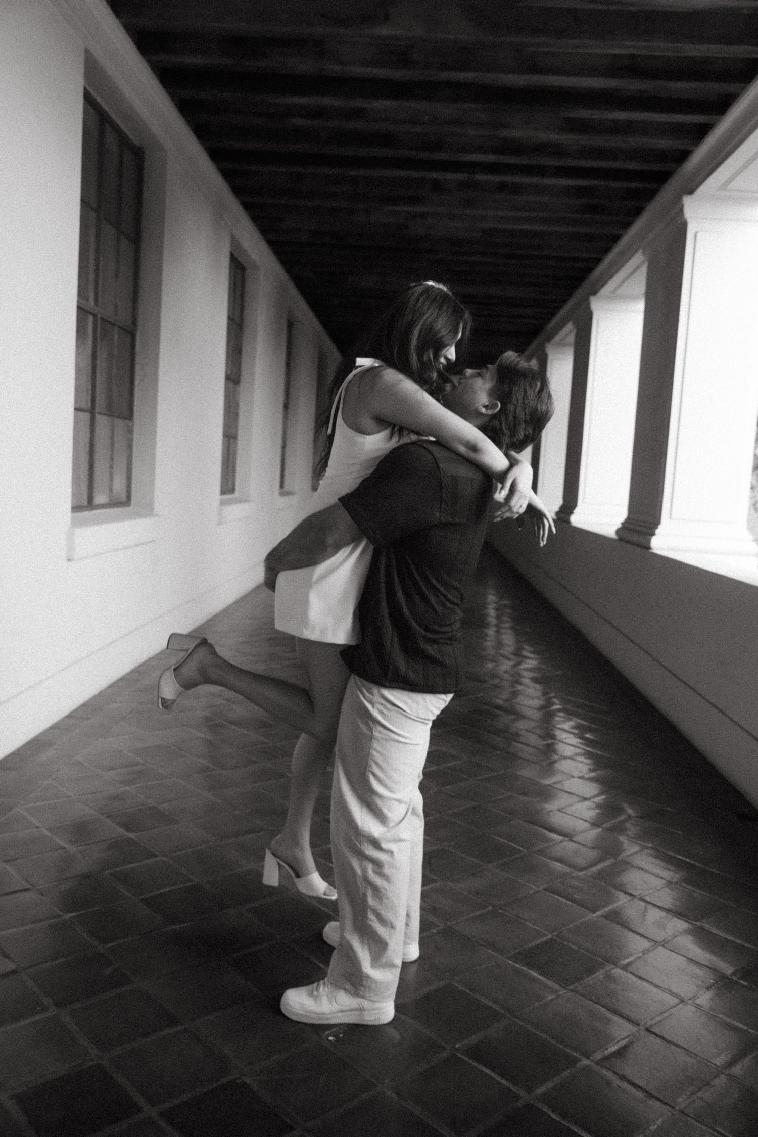 Black and white photo of a couple sharing a romantic embrace and kiss in a long corridor.