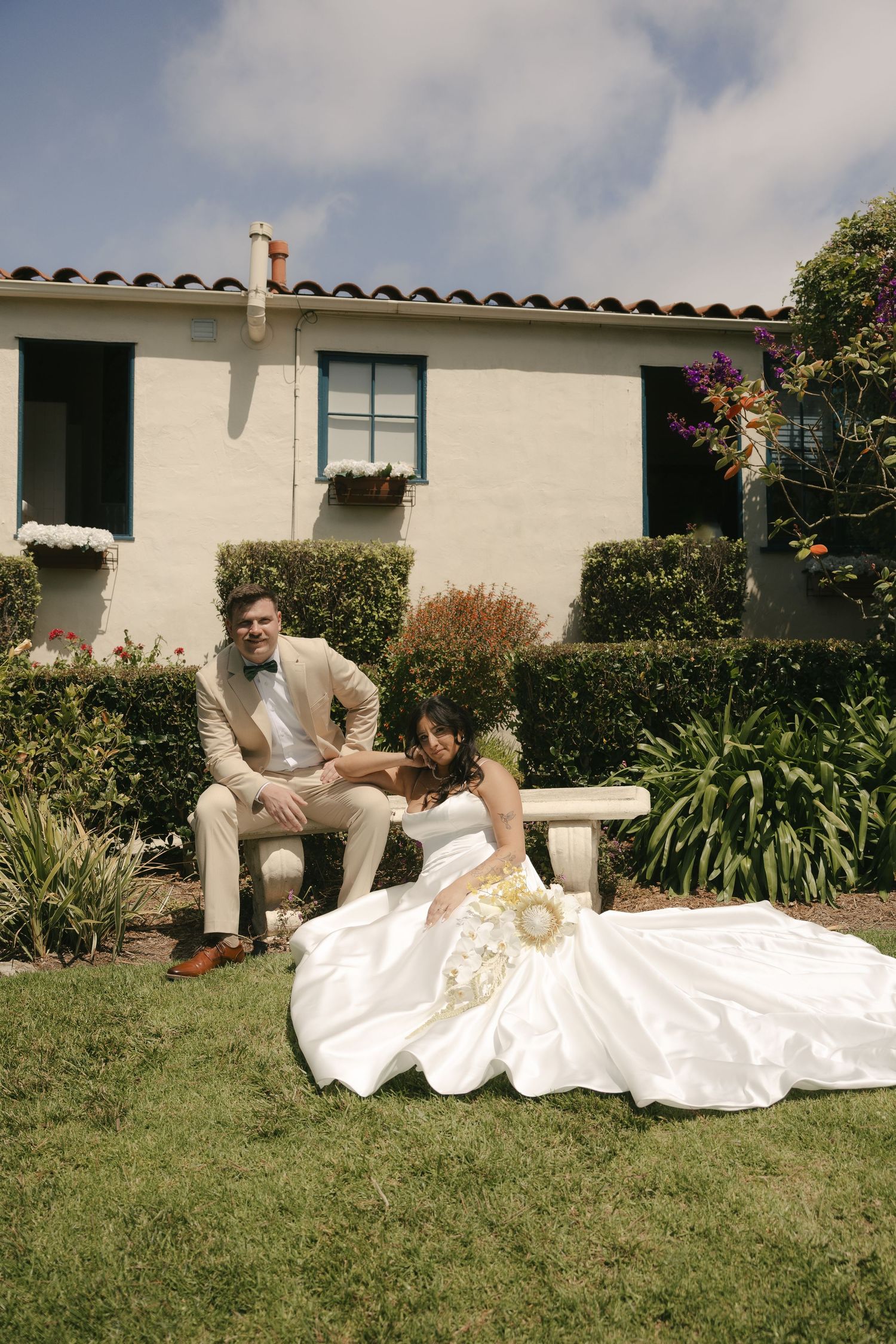 Bride in voluminous white gown sits on garden bench with groom in tan suit outside a Spanish-style villa.