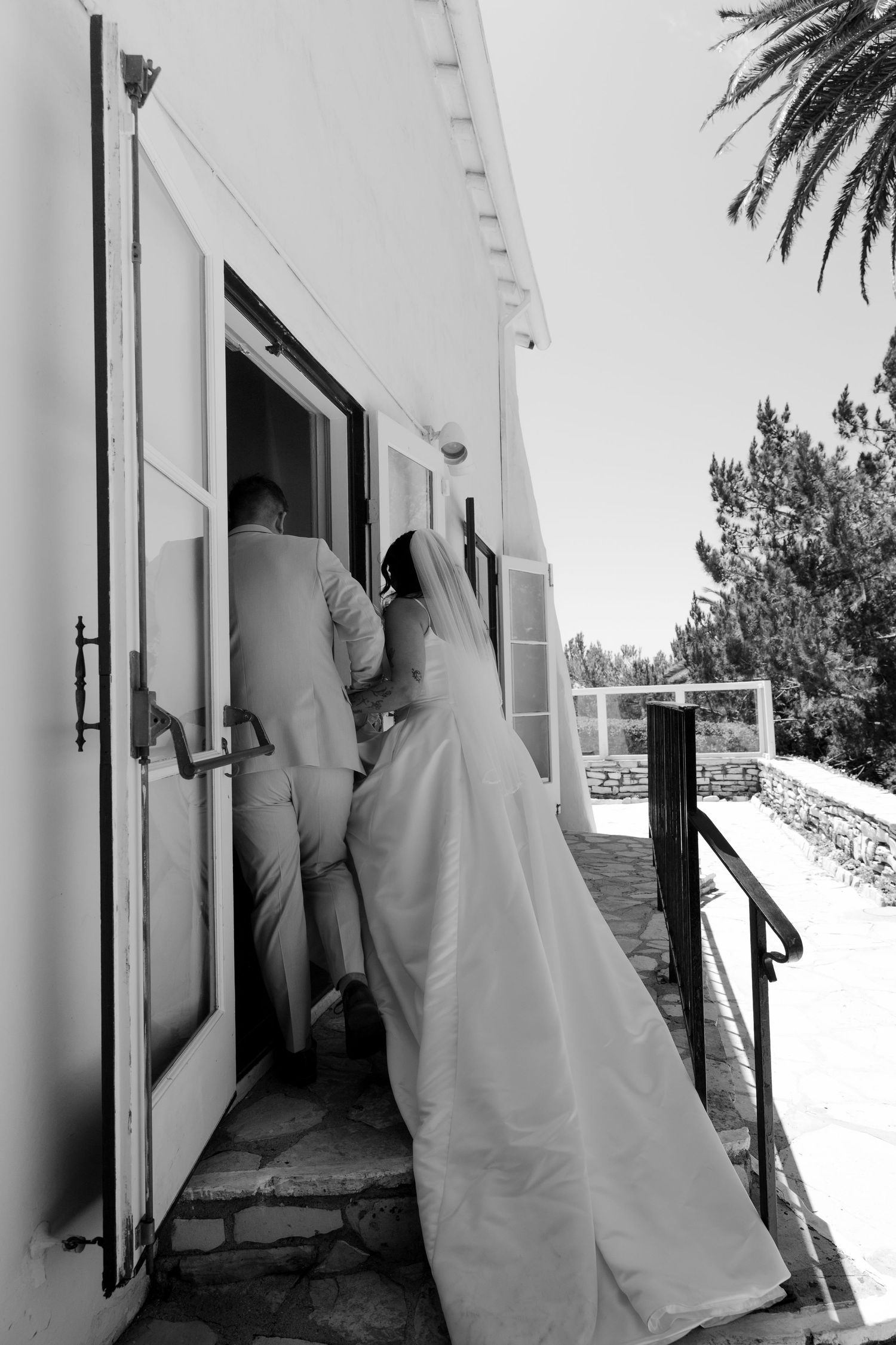 Black and white photo of a bride in a flowing gown entering a white chapel with groom on a sunny wedding day.