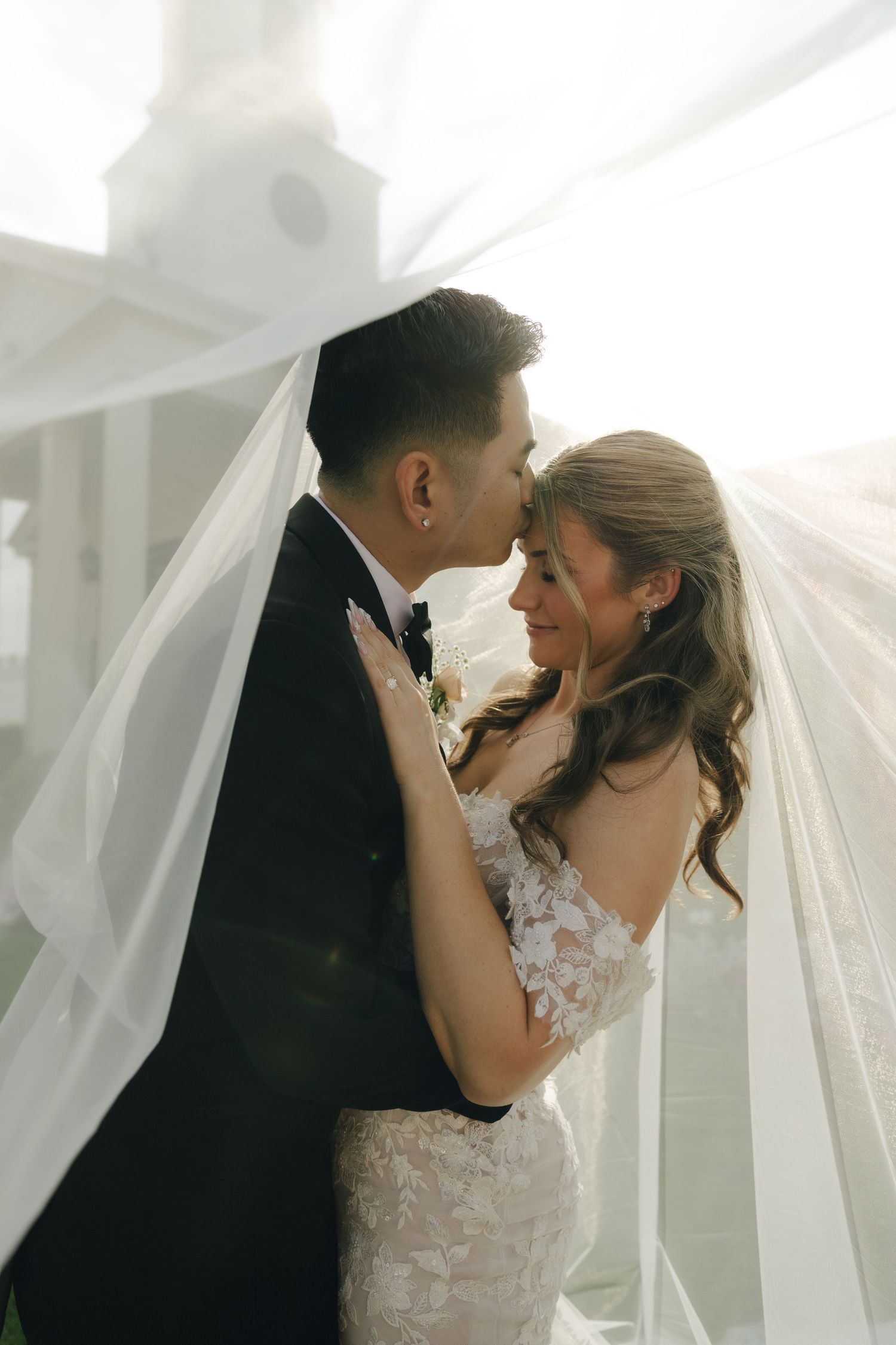 Bride and groom share an intimate moment under flowing white veil with sunlit church in background.