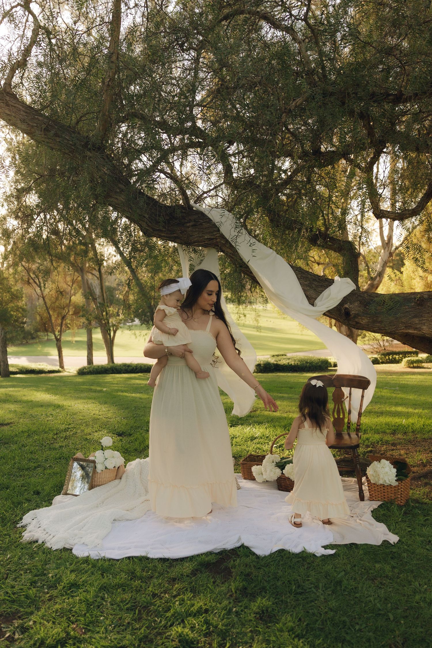 Woman in white gown holds baby while young girl in floral crown reaches up under a sunlit tree outdoors.