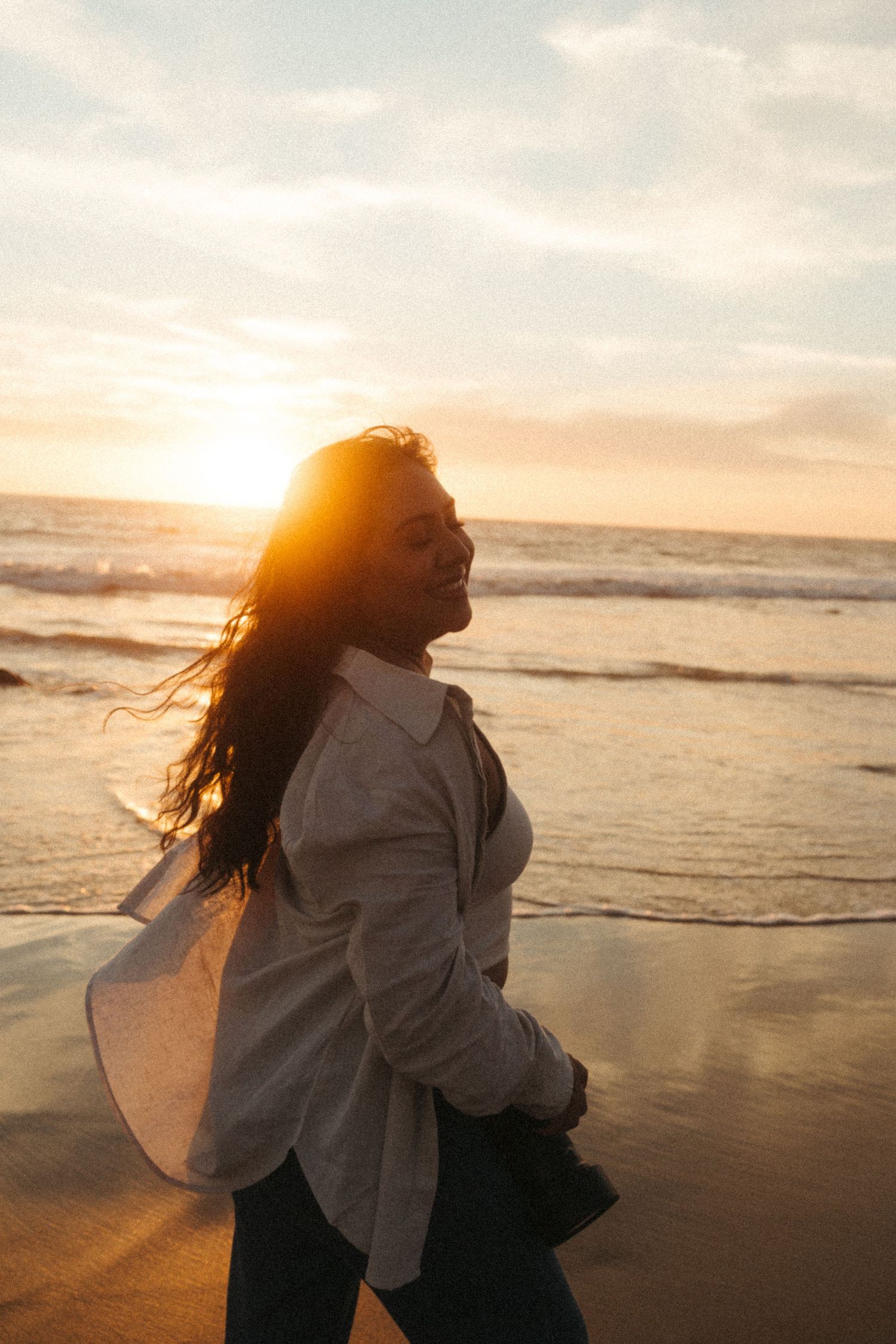 Woman smiling on beach at golden sunset, hair blowing in wind, wearing oversized light gray shirt.
