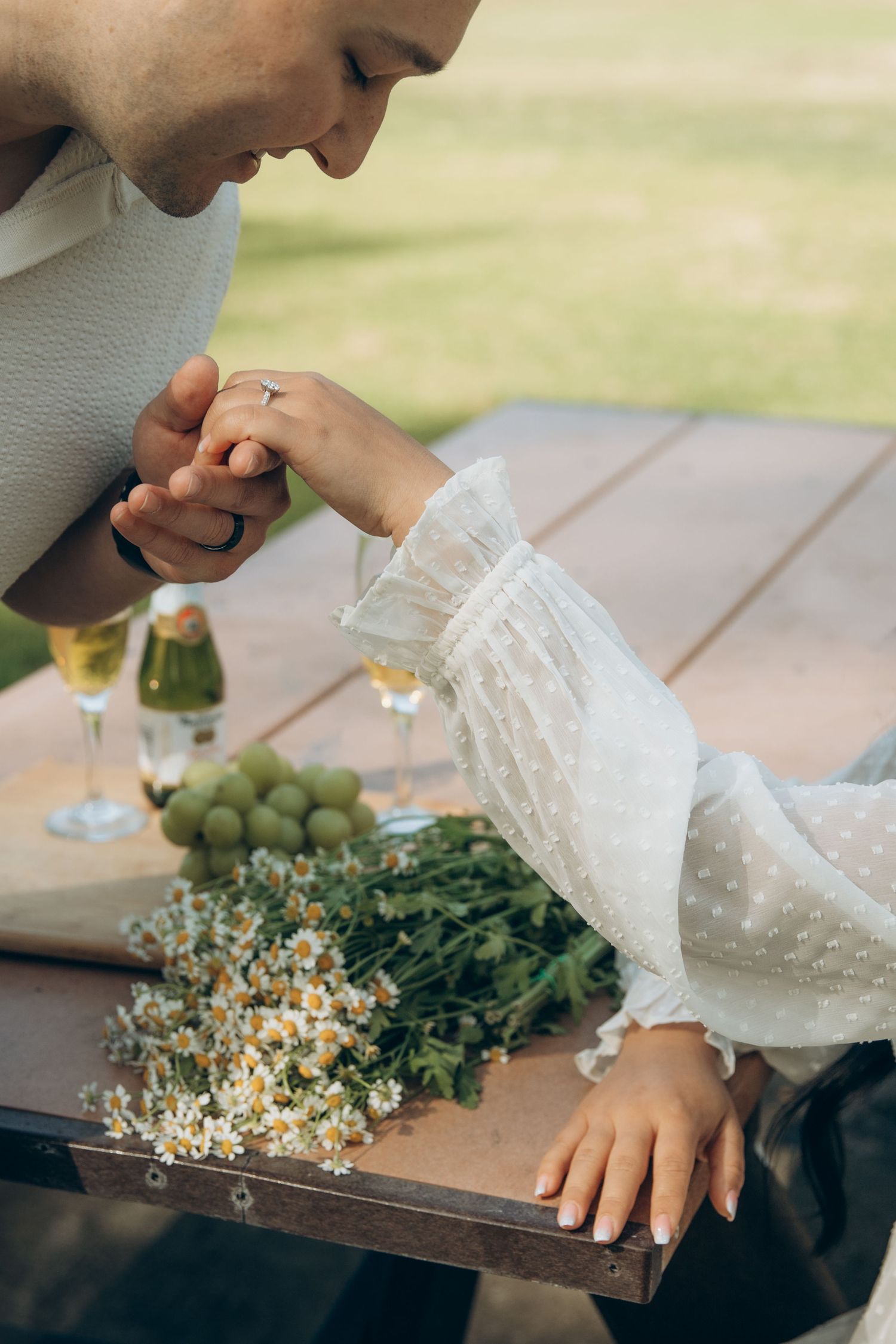 Man kissing womans hand at a picnic table with champagne, grapes, and daisy bouquet in a garden setting.