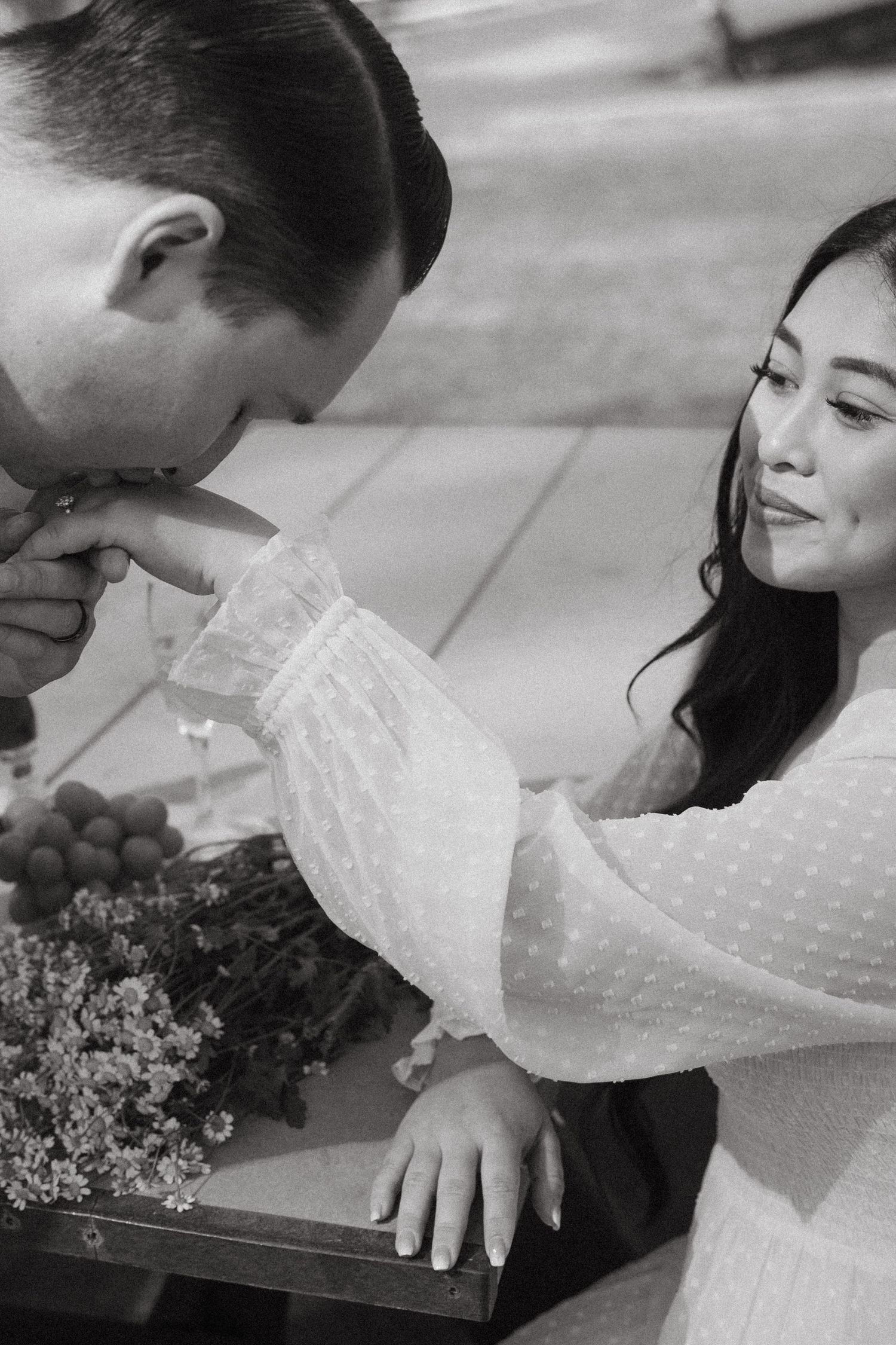 Black and white photo of man kissing womans hand at a romantic outdoor picnic with flowers and grapes.