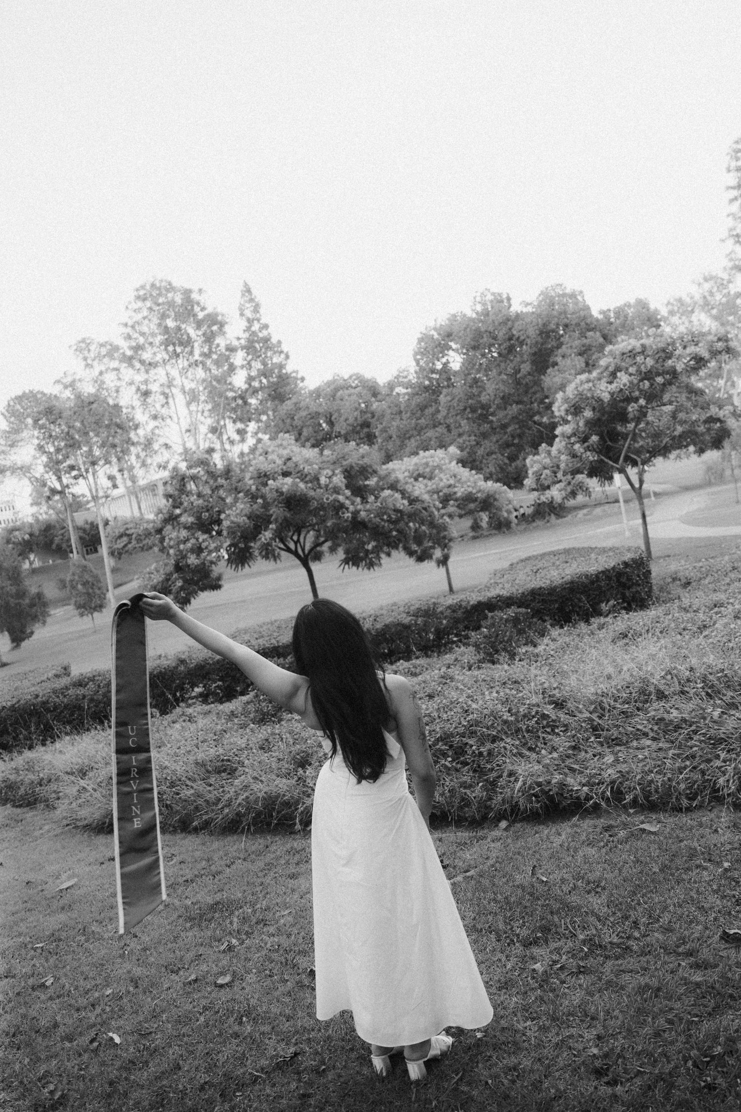 Black and white photo of woman in white dress holding graduation sash outstretched in park setting.