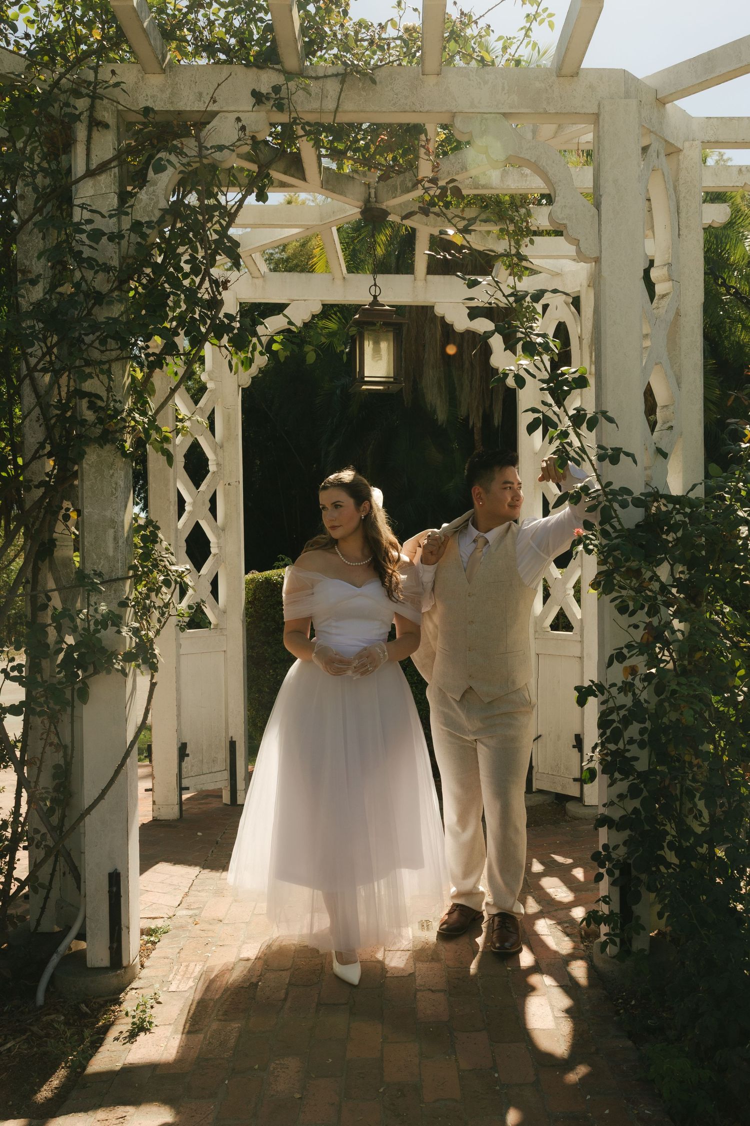 Bride and groom standing together under a white garden arbor draped with lush green foliage.