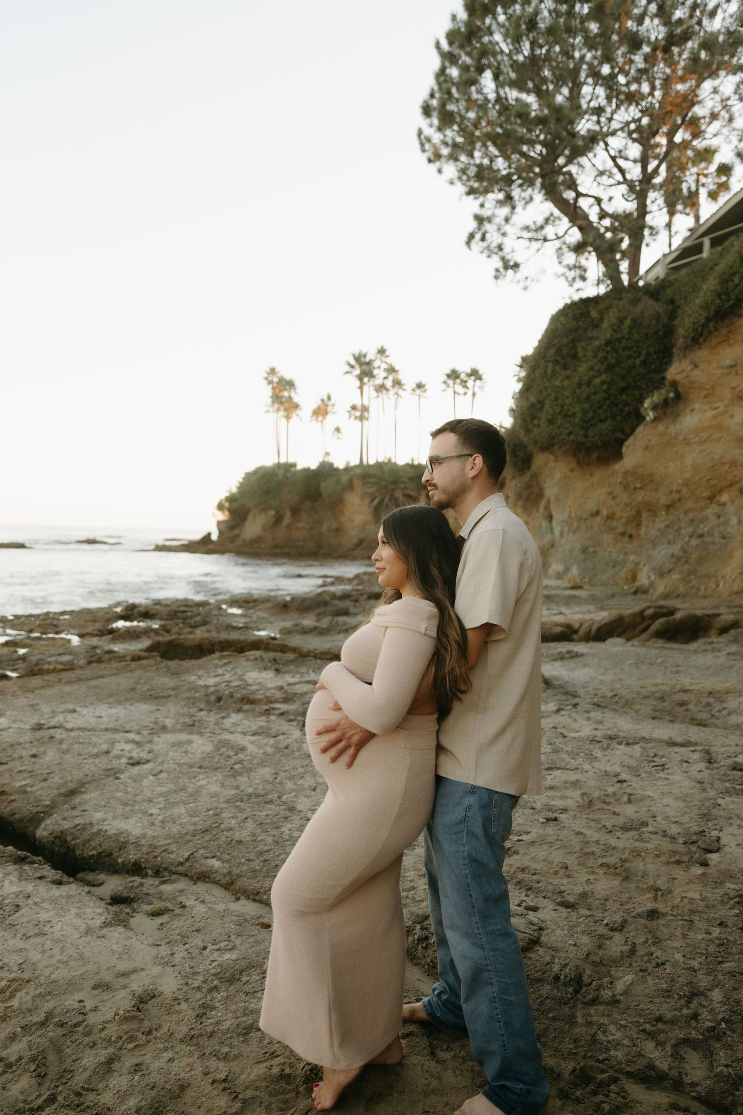 Pregnant couple embraces on rocky beach at sunset with palm trees and cliffs in background.