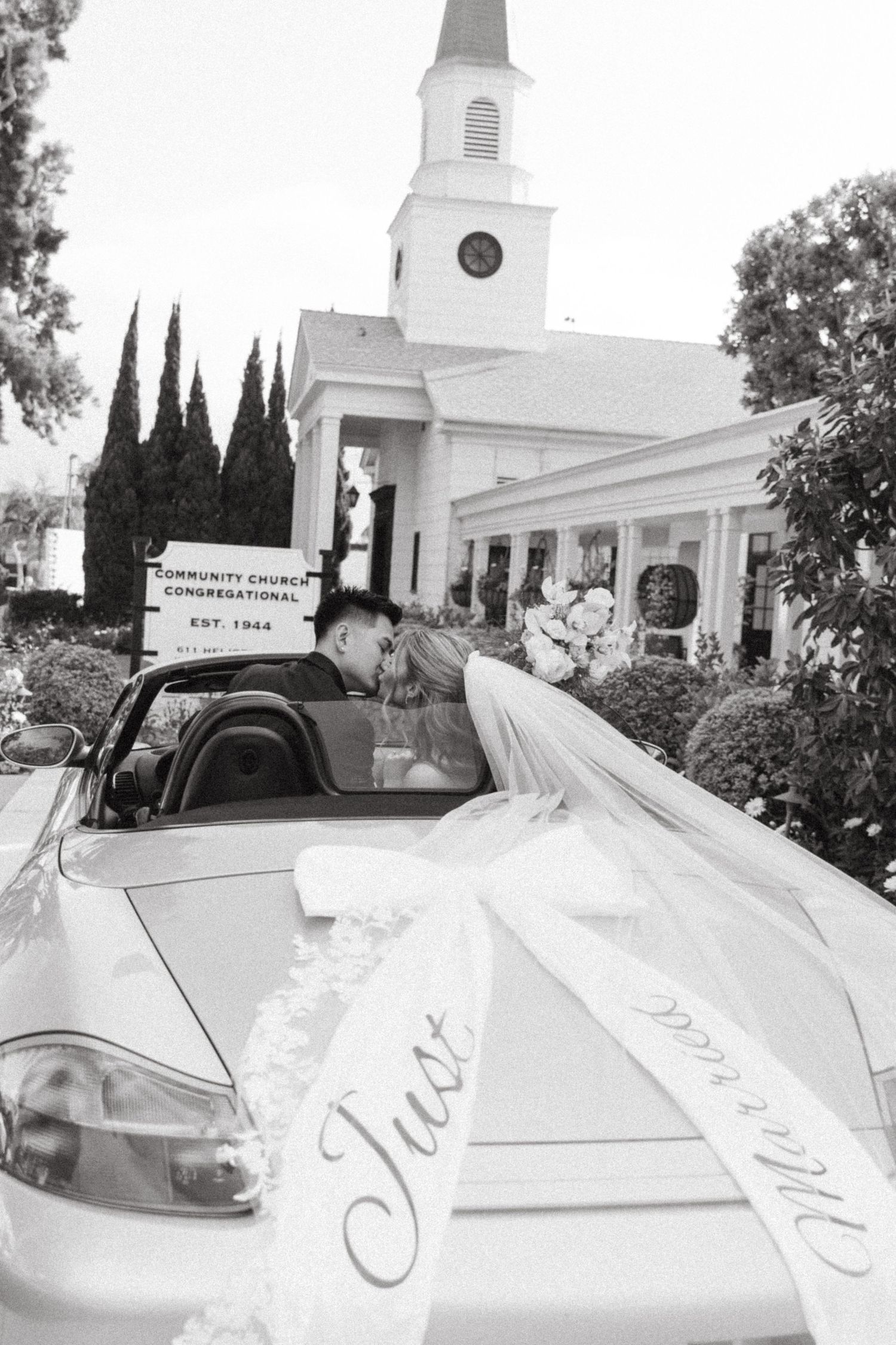 Black and white photo of newlyweds kissing in convertible car marked Just Married outside church.