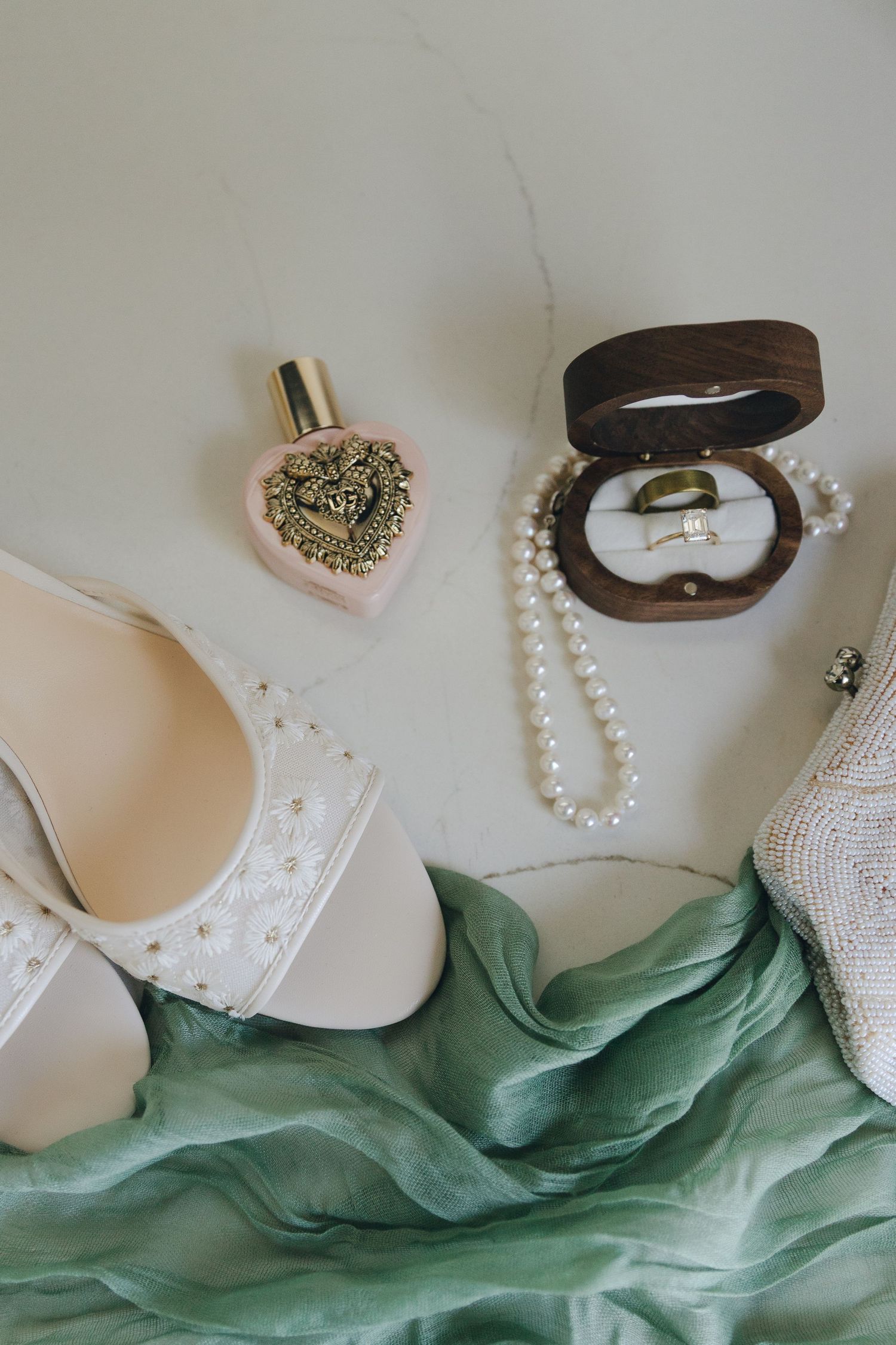 Bridal flat lay featuring pearl heels, perfume, ring box with wedding bands, and green silk ribbon.