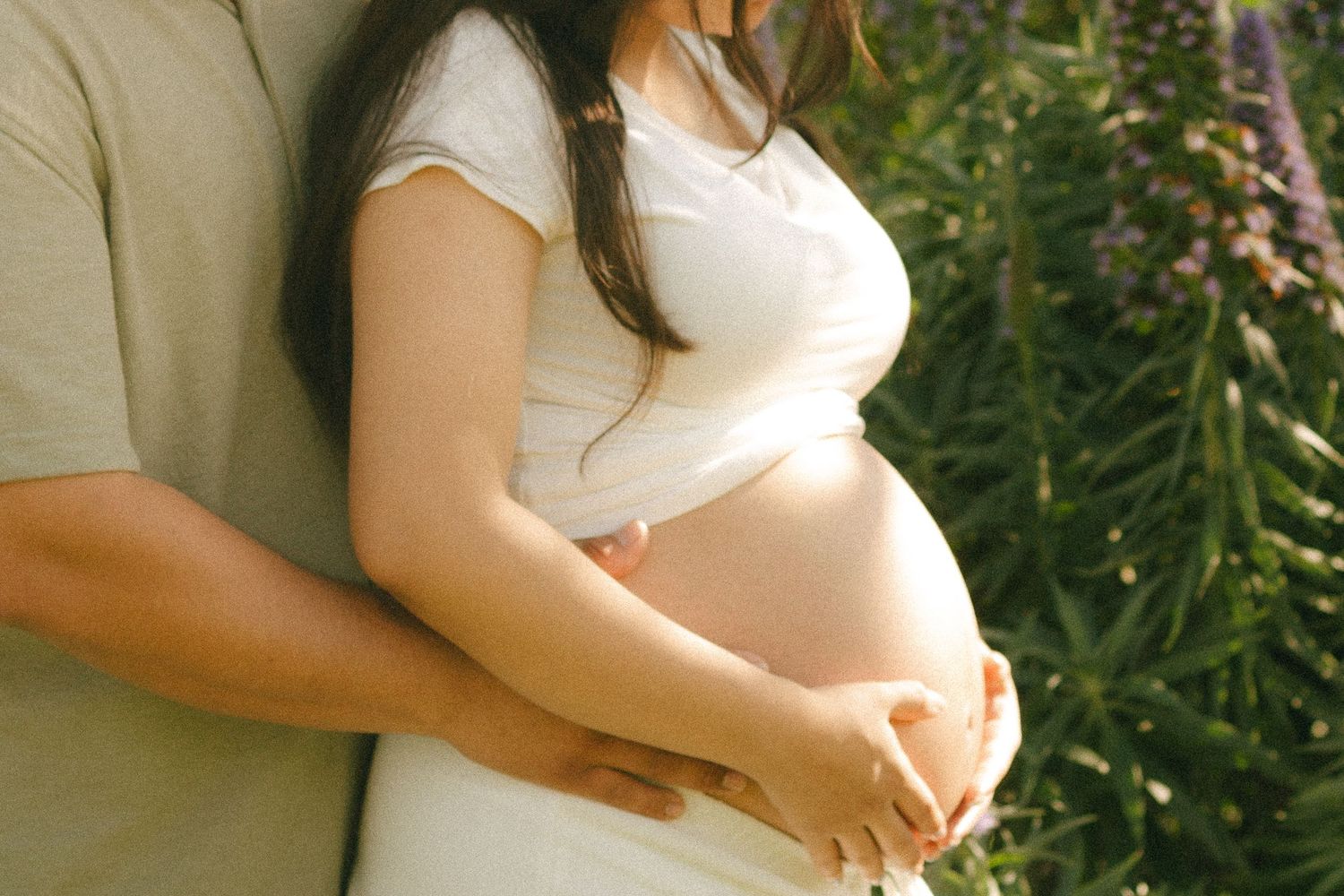 Couple sharing tender moment outdoors, man embracing pregnant woman in white dress among purple wildflowers.