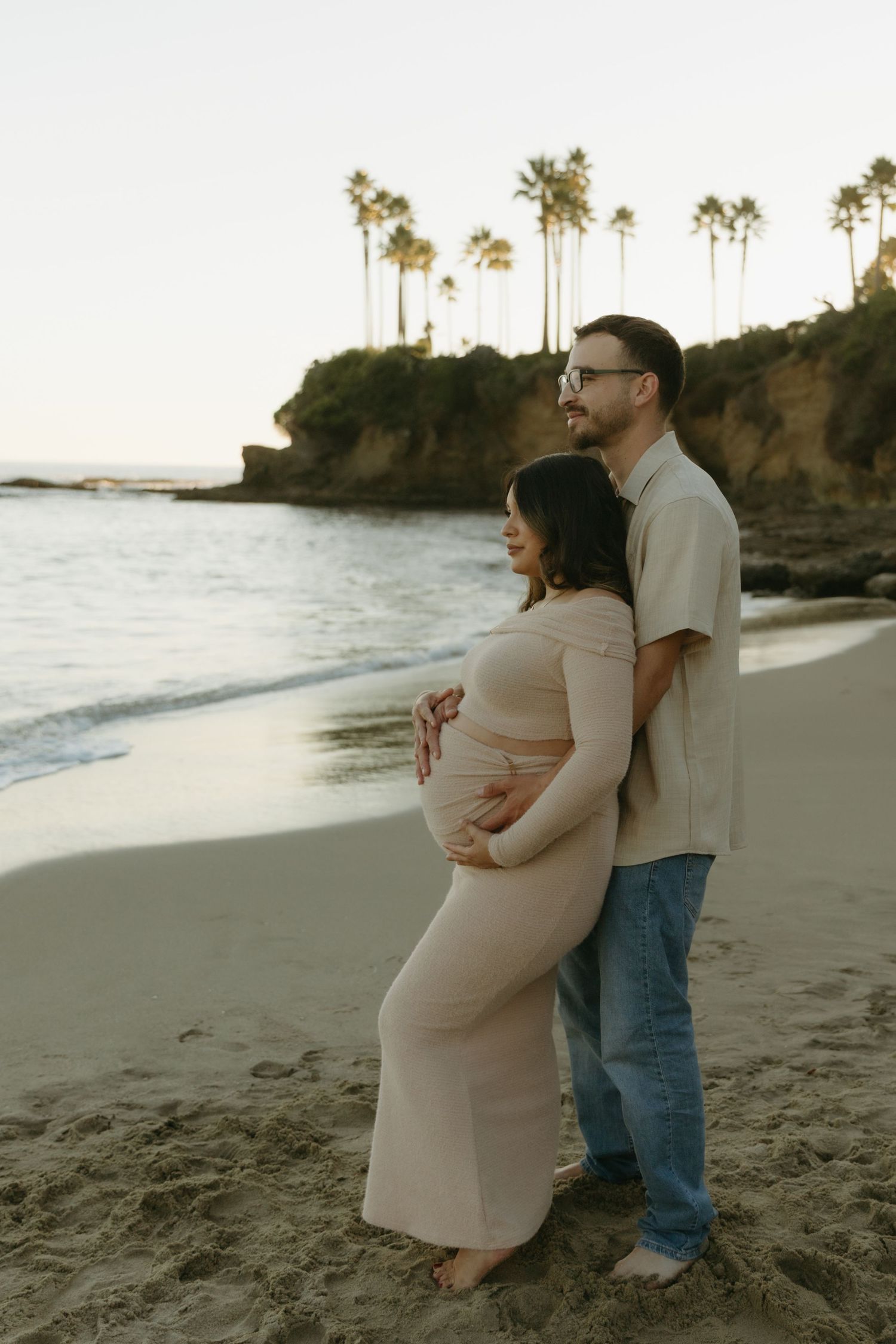 Couple stands together on sandy beach at sunset, man embracing pregnant woman from behind near ocean.