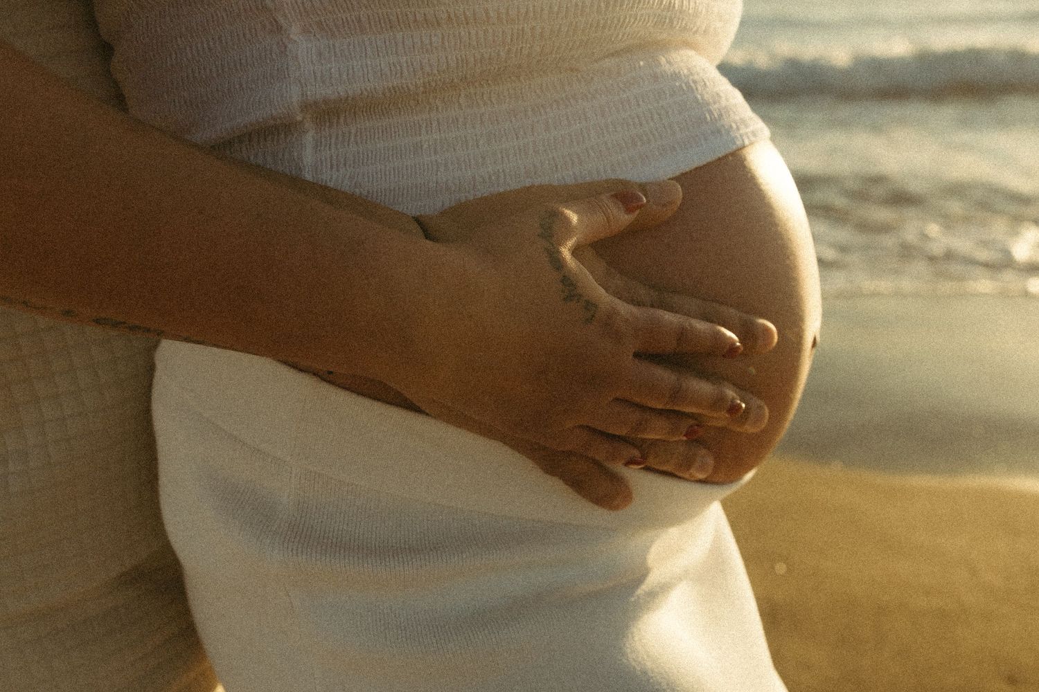 A pregnant woman in white dress cradles her baby bump on a sunny beach by the ocean.