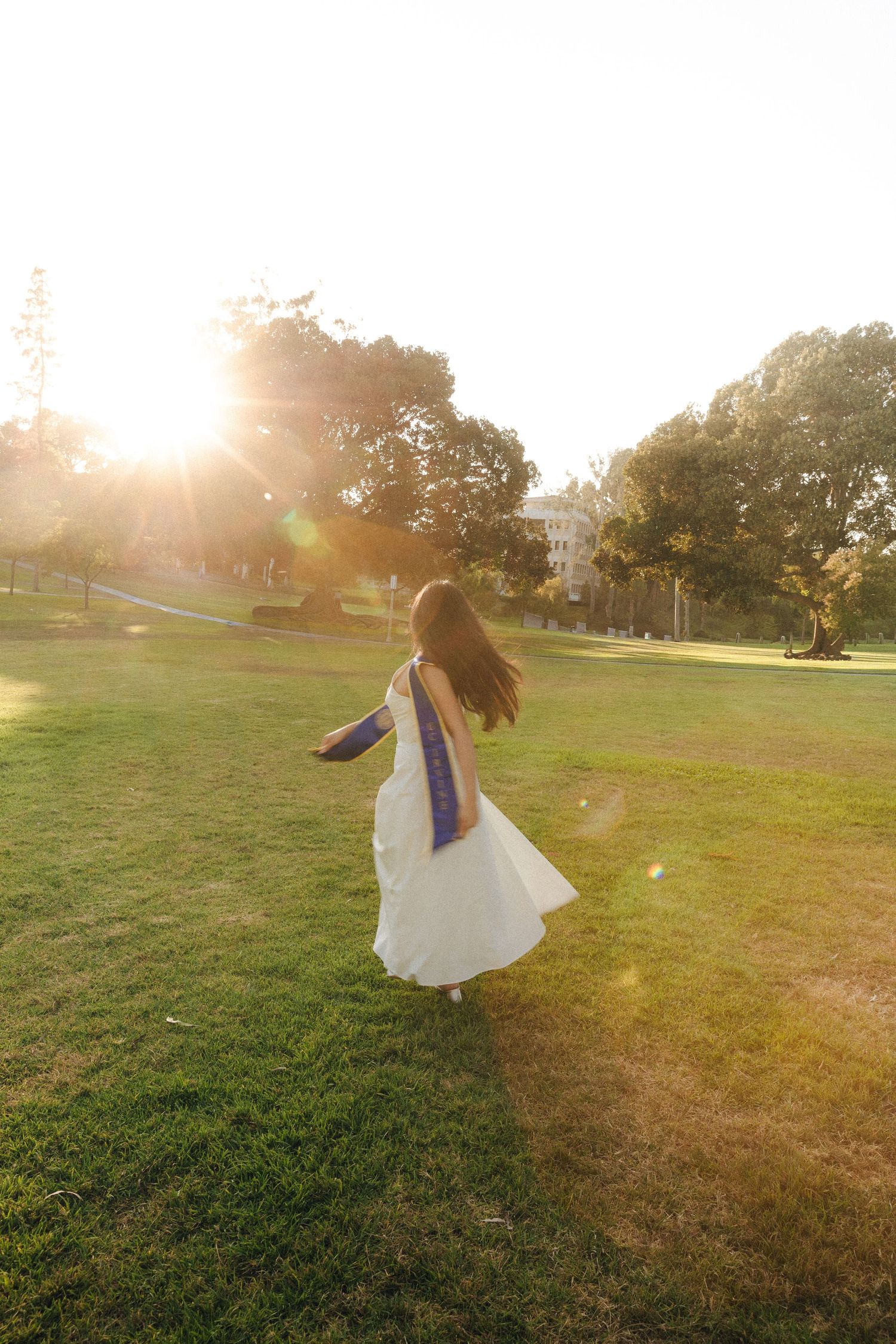 Woman in white dress and blue graduation sash twirling on green grass during golden hour sunset.