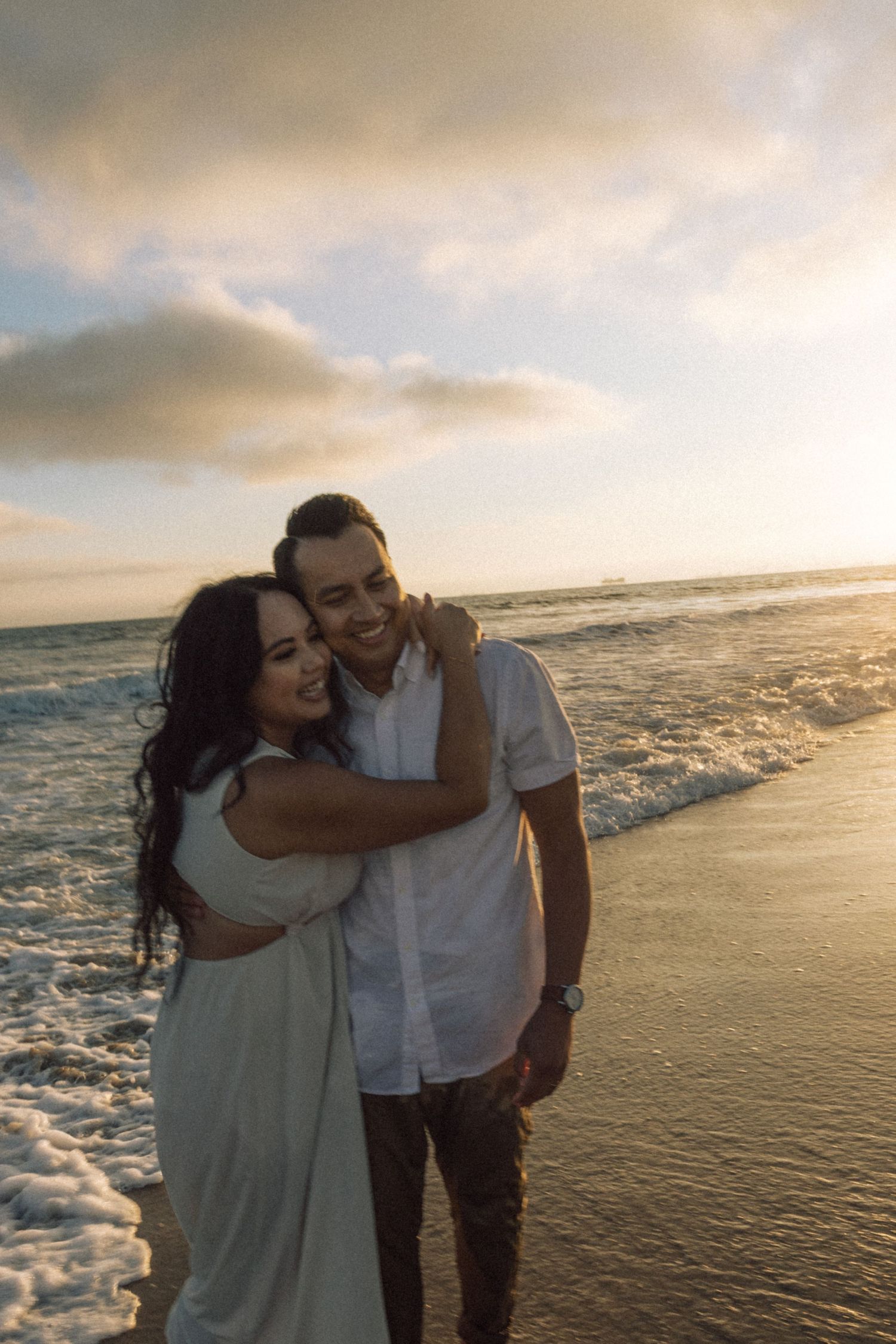 Happy couple embracing on a beach at golden hour with ocean waves and dramatic sunset sky.